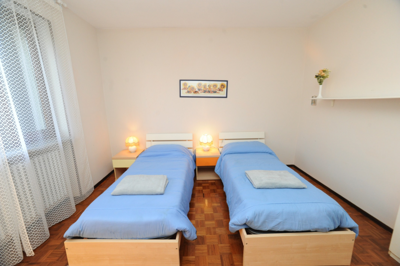 Due letti con lenzuola blu in una stanza con pareti bianche, una finestra, piccoli comodini e un piccolo quadro sul muro.