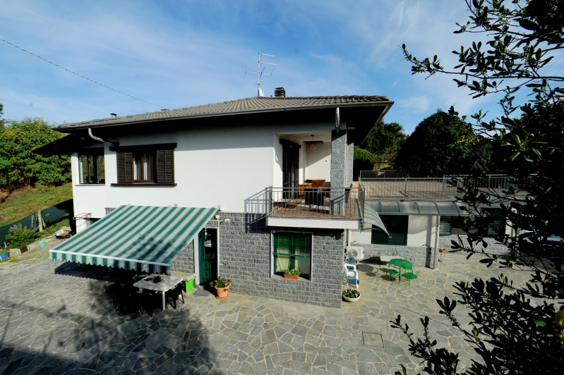 Casa a due piani con tenda da sole verde, facciata in pietra, balcone e patio. Giornata di sole, cielo azzurro.