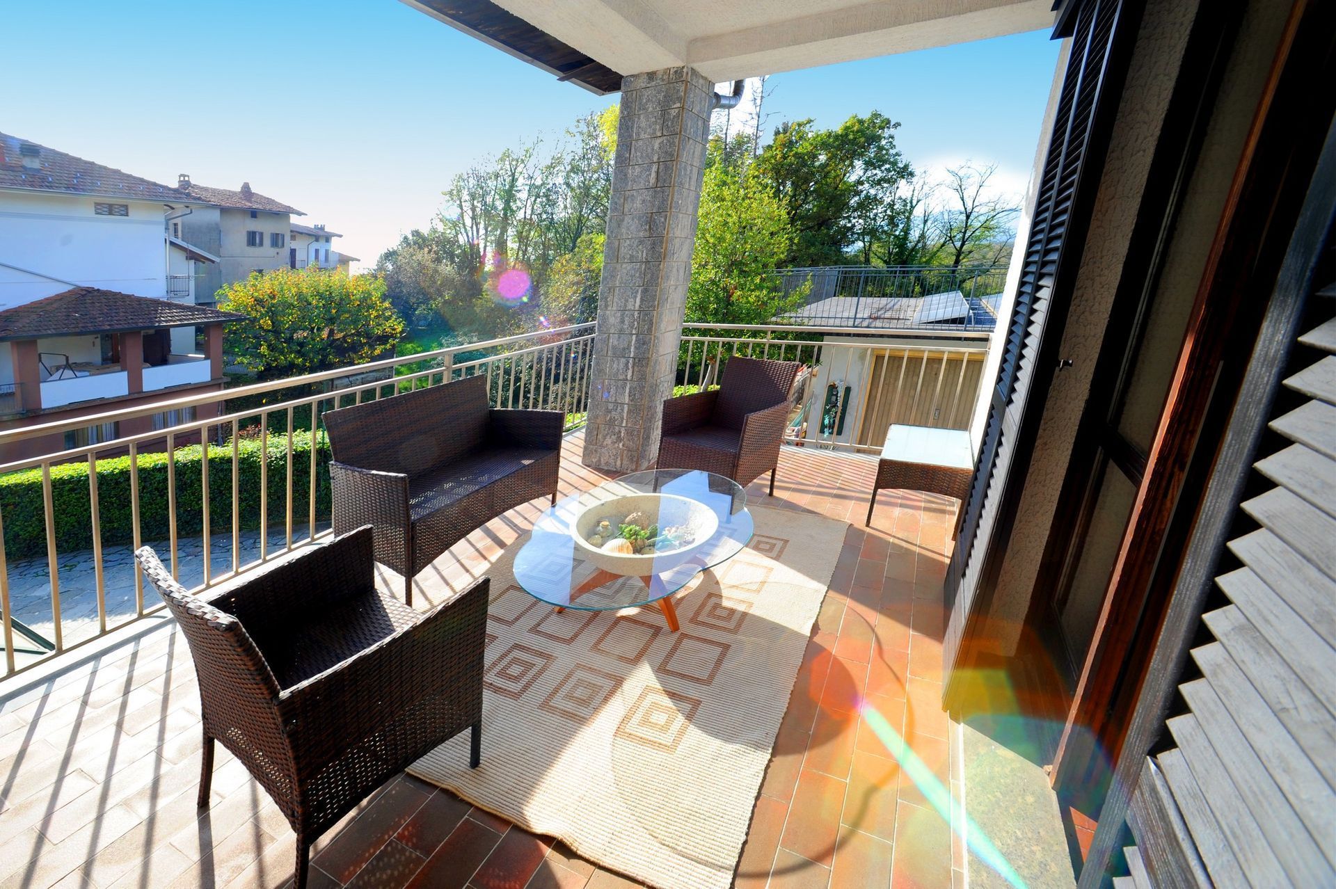 Balcone con mobili in vimini: divano, sedie, tavolo, tappeto. Si affaccia su una zona residenziale alberata.