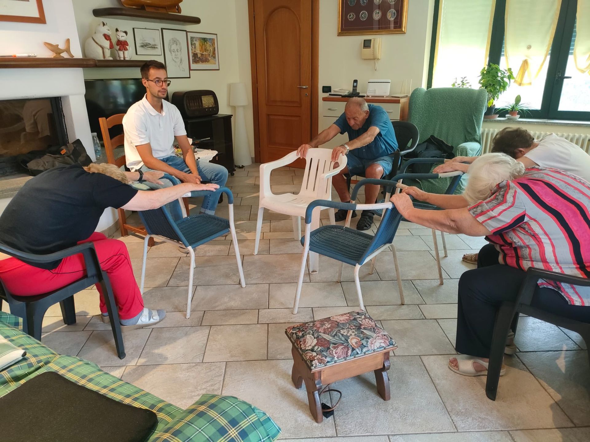 Persone che si stiracchiano sulle sedie, guidate da un uomo in camicia bianca. All'interno, con camino e finestre.