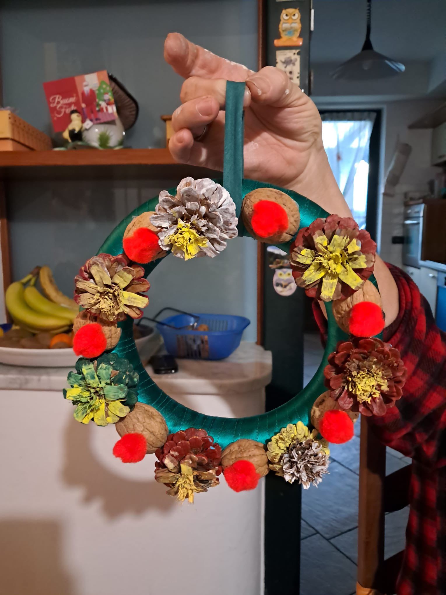 Mano che regge una ghirlanda natalizia verde acqua decorata con pigne, pompon rossi e nastro, in casa.