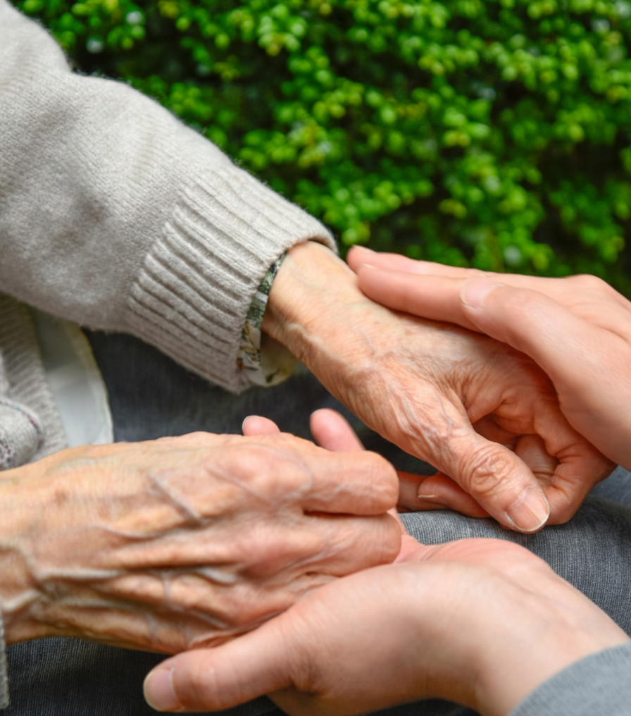 Due mani, una anziana e una più giovane, che si tengono delicatamente. Sullo sfondo, fogliame verde.