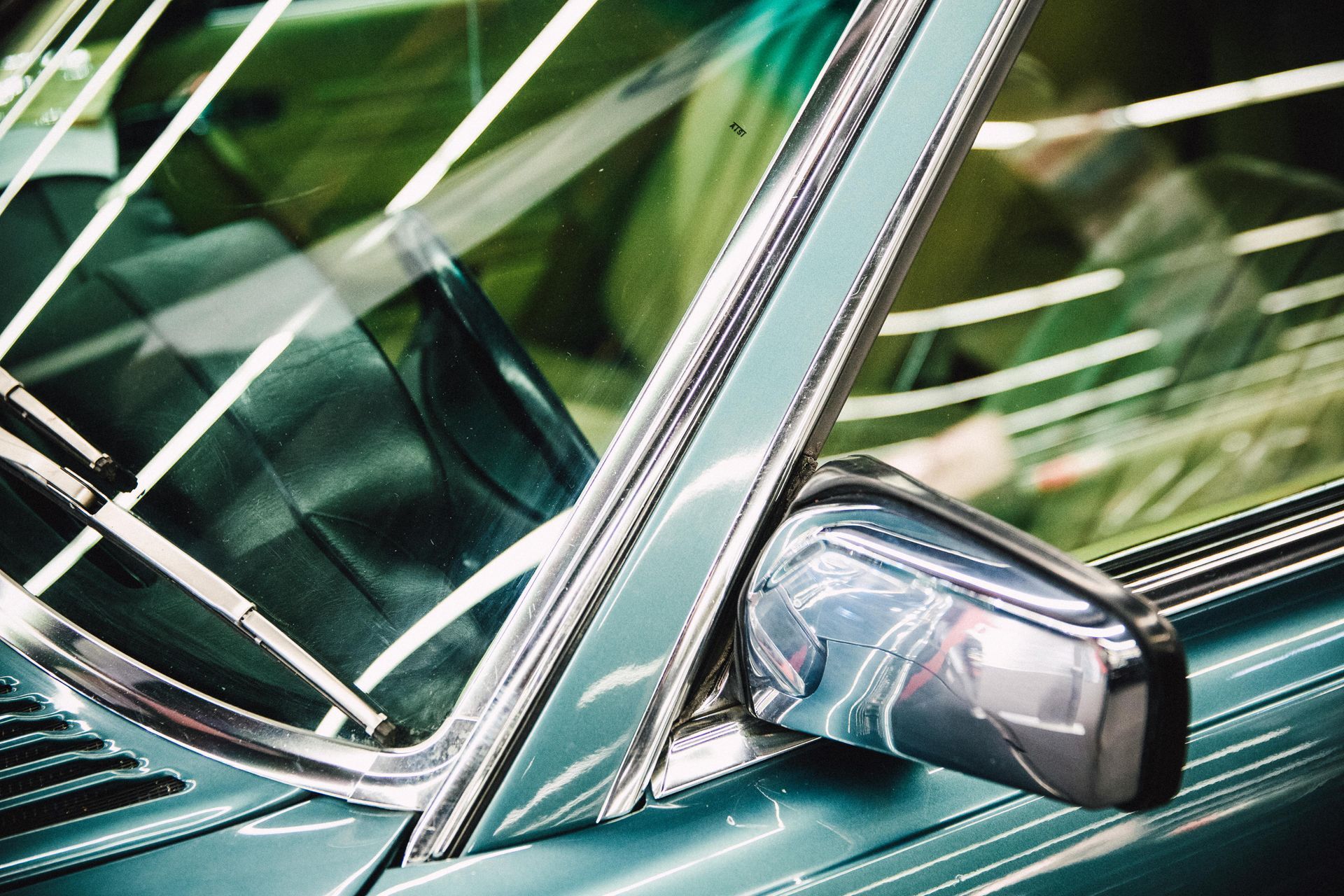 Close-up of a teal classic car's side window, chrome trim, and side mirror reflecting surroundings.