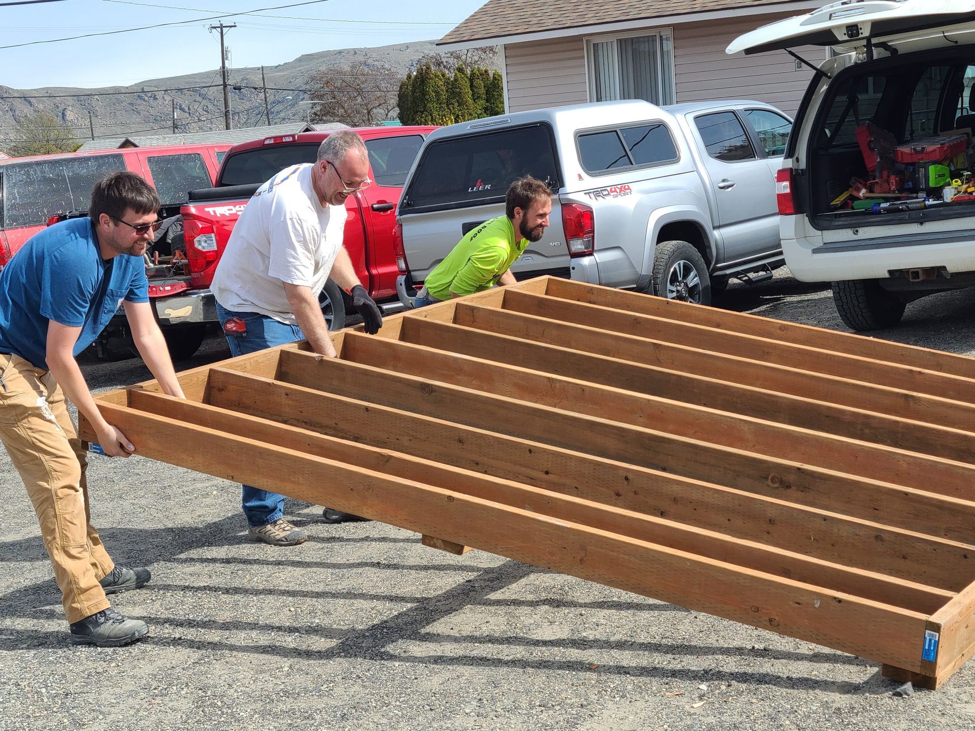 A group of three men raise a large piece of wood in a parking lot.