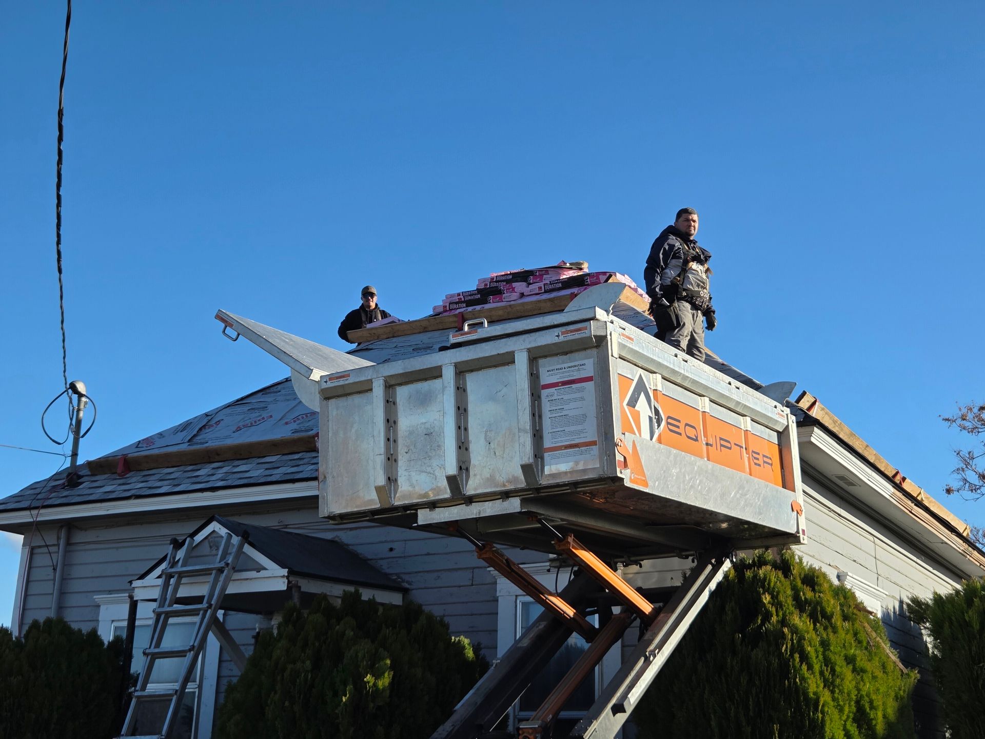 Two workers on a rooftop, using a lift to transport roofing materials against a clear blue sky.