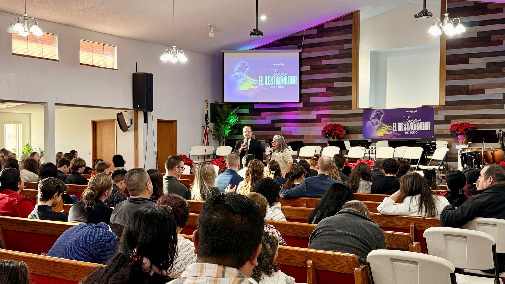People sit in pews in a church hall, facing a stage with a projection screen and two speakers at a podium.