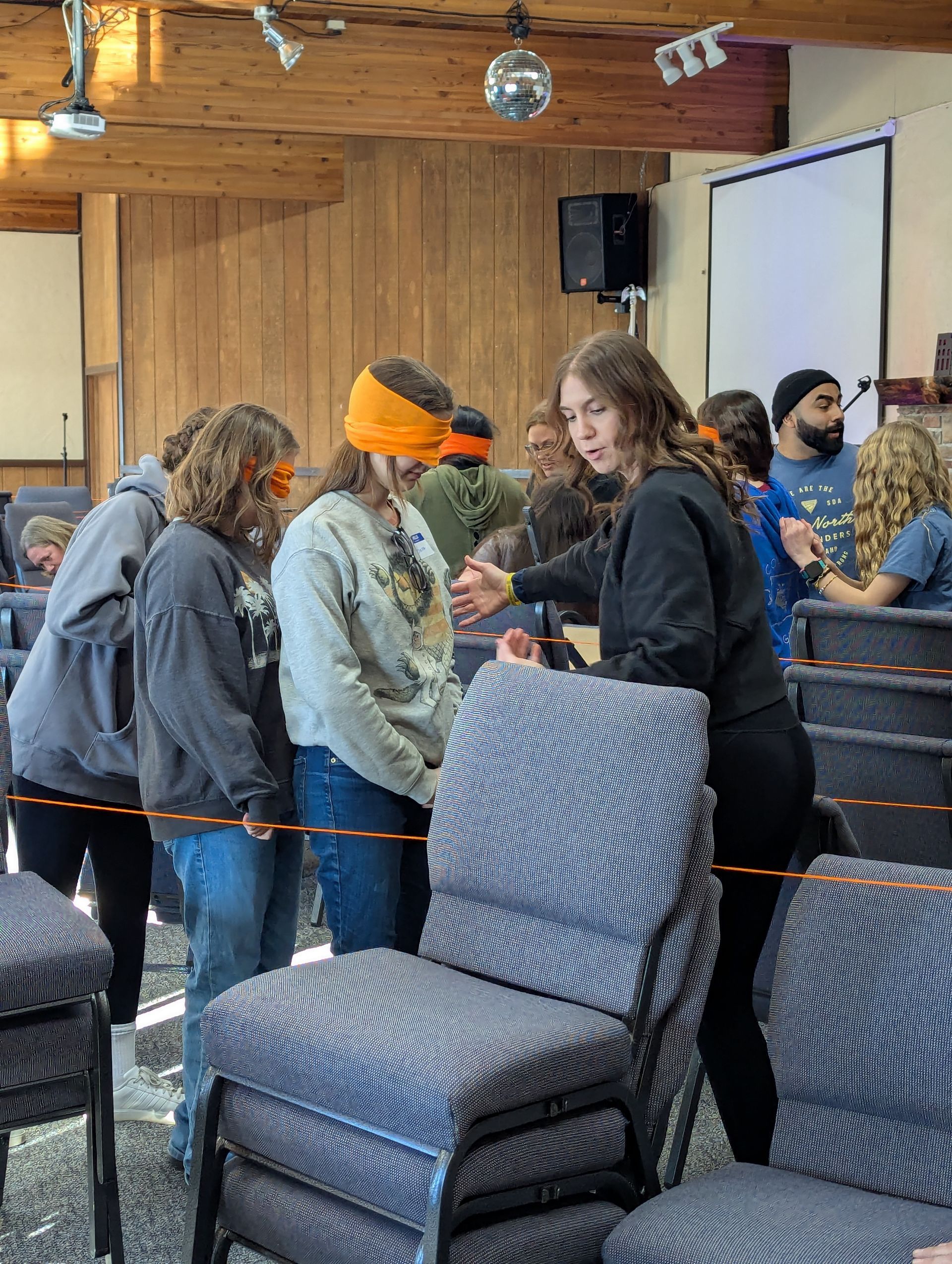 A blindfolded teen trusts their fellow pathfinders to find a path for them through a maze of chairs.