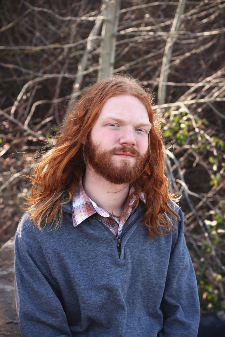 Asher Peterson, a man with long red hair and a beard is wearing a gray sweater.