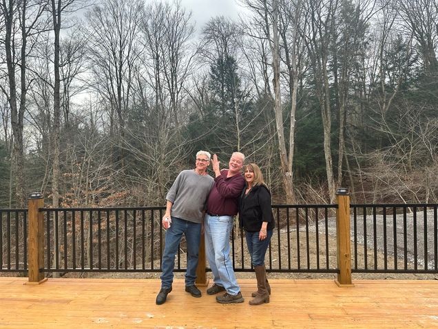 Three people are posing for a picture on a deck with trees in the background.