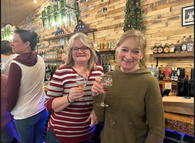 Two women are standing next to each other in a bar holding wine glasses.
