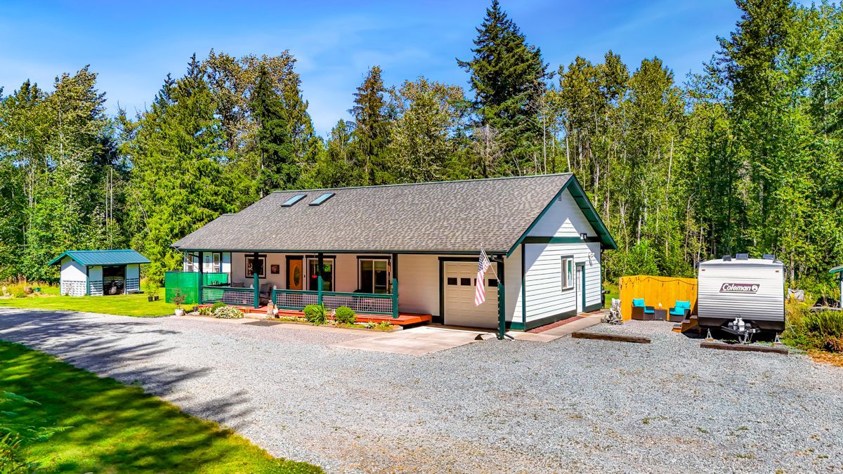Charming single-story home with covered front porch, skylights, and attached garage, set on a spacious gravel drive surrounded by lush forest and mature trees.