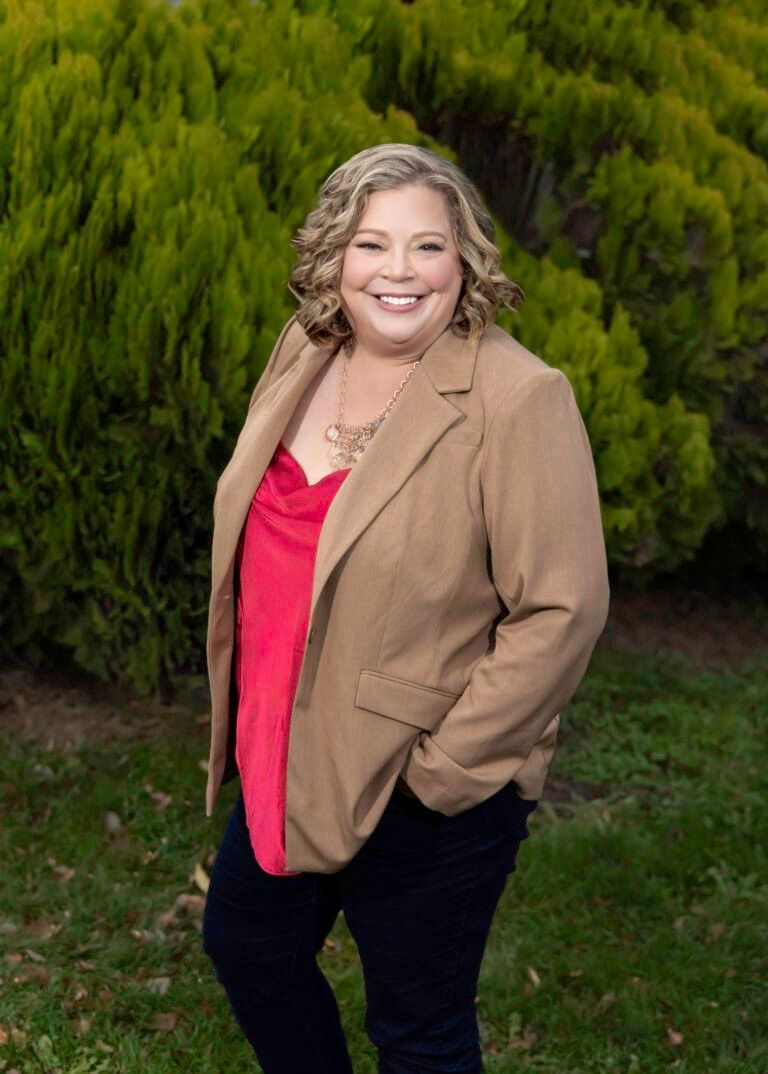 Andi Dyer in a tan jacket and a red top is standing in the grass.