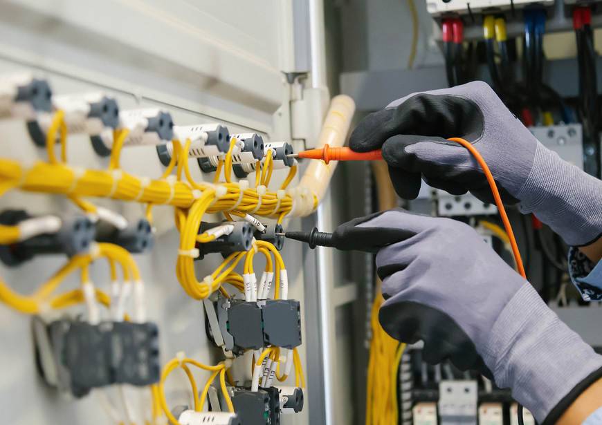Gloved hands use a multimeter probe to test electrical connections on a control panel with organized yellow wiring.