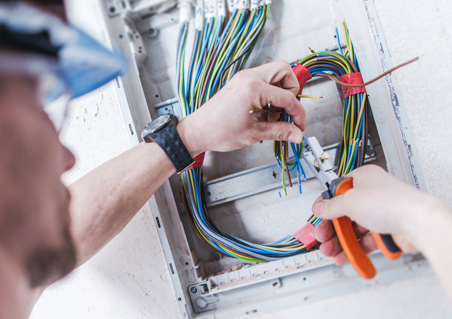 Electrician with safety glasses works on colored wires inside an electrical panel using wire stripping pliers.