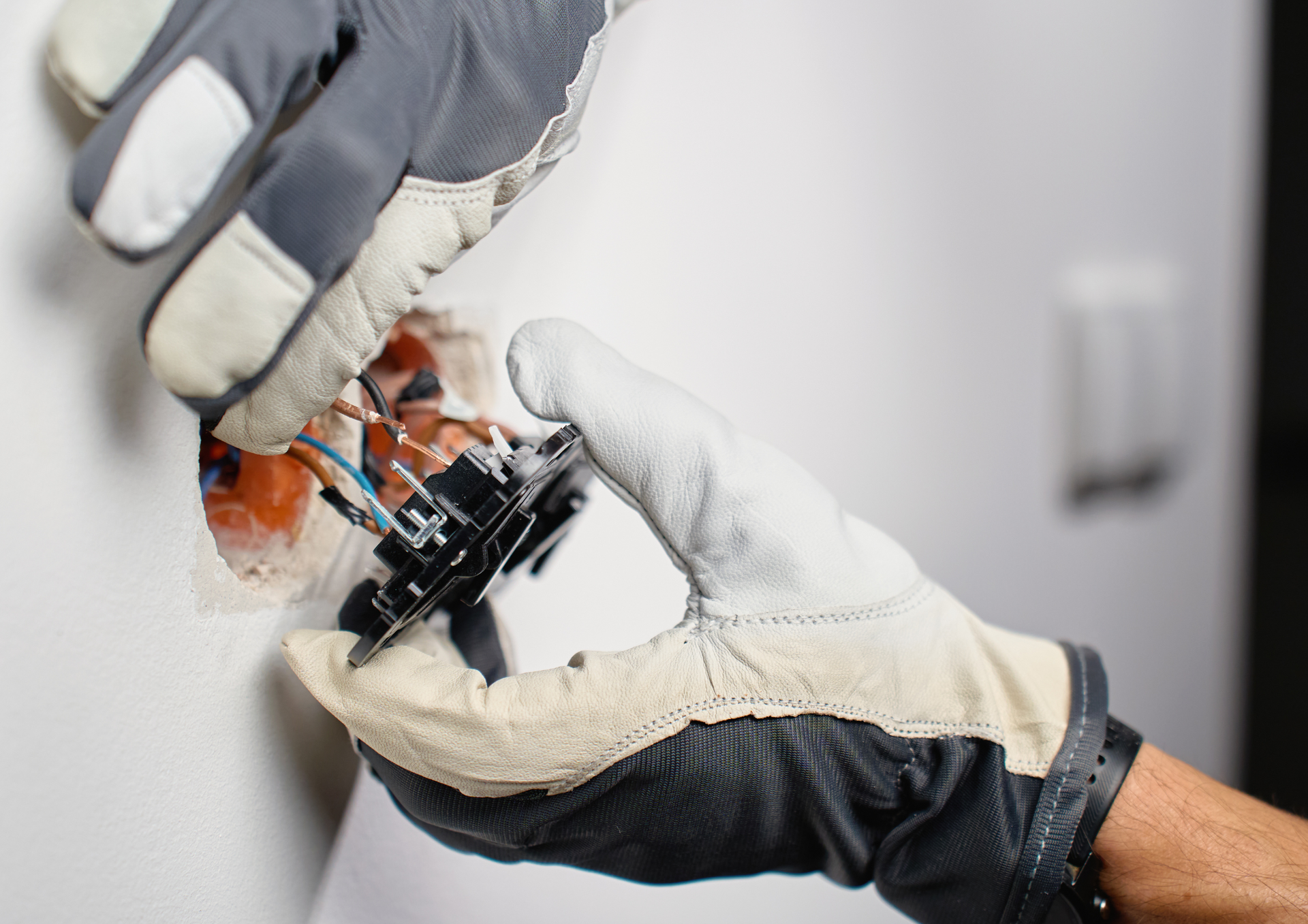 A pair of gloved hands works to install or repair a wall-mounted electrical outlet.