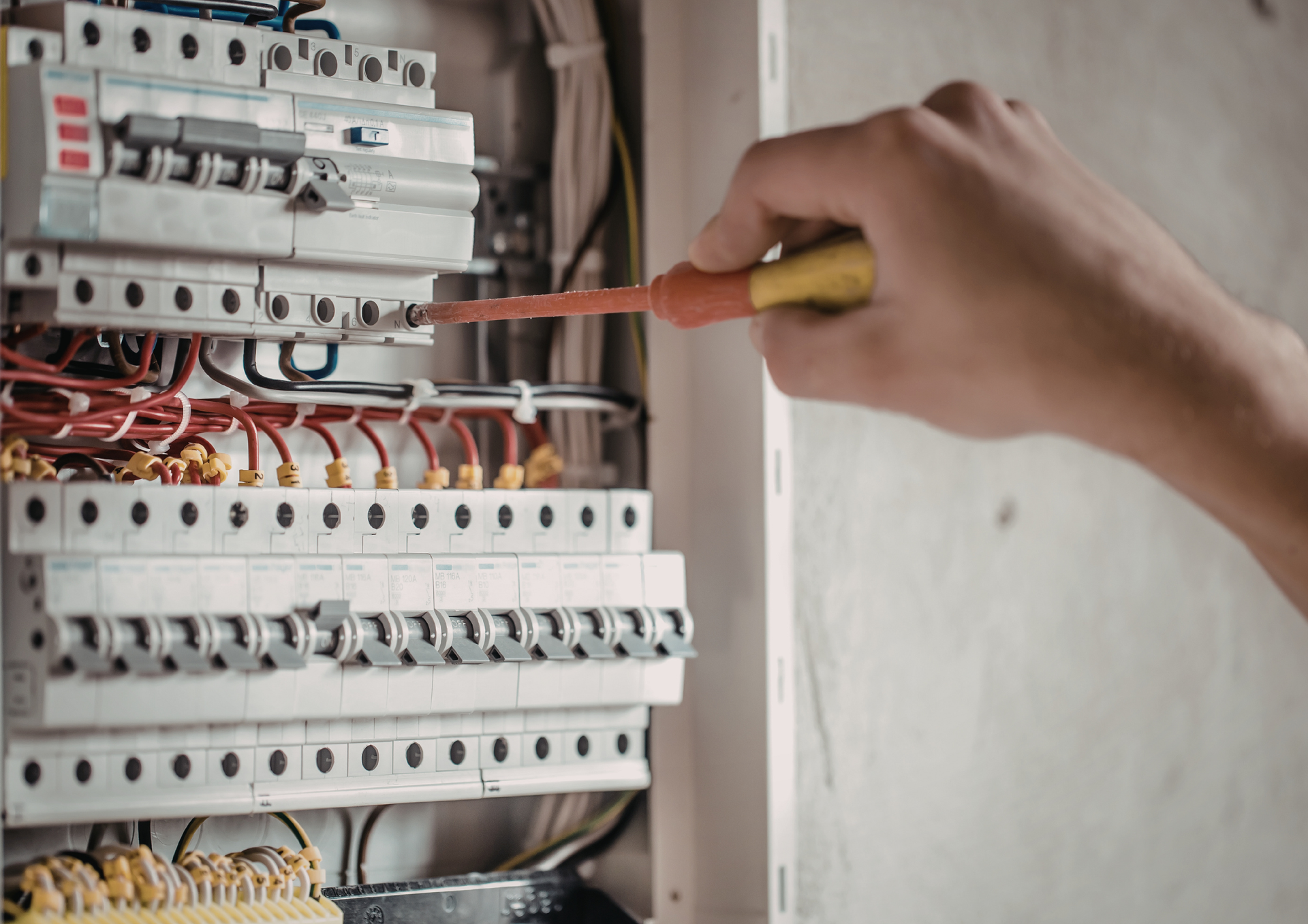A hand uses a yellow and orange screwdriver to tighten a screw on an electrical circuit breaker panel.