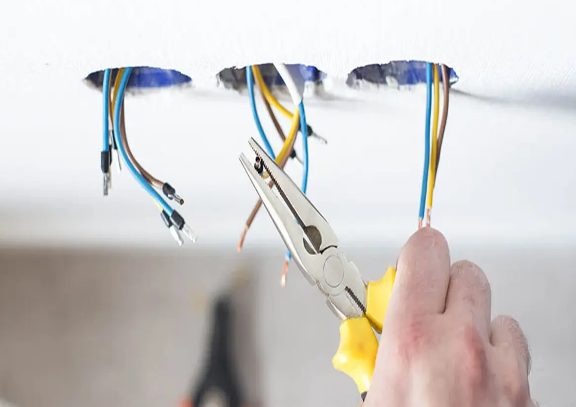A hand uses needle-nose pliers to strip electrical wires hanging from three holes in a white ceiling.