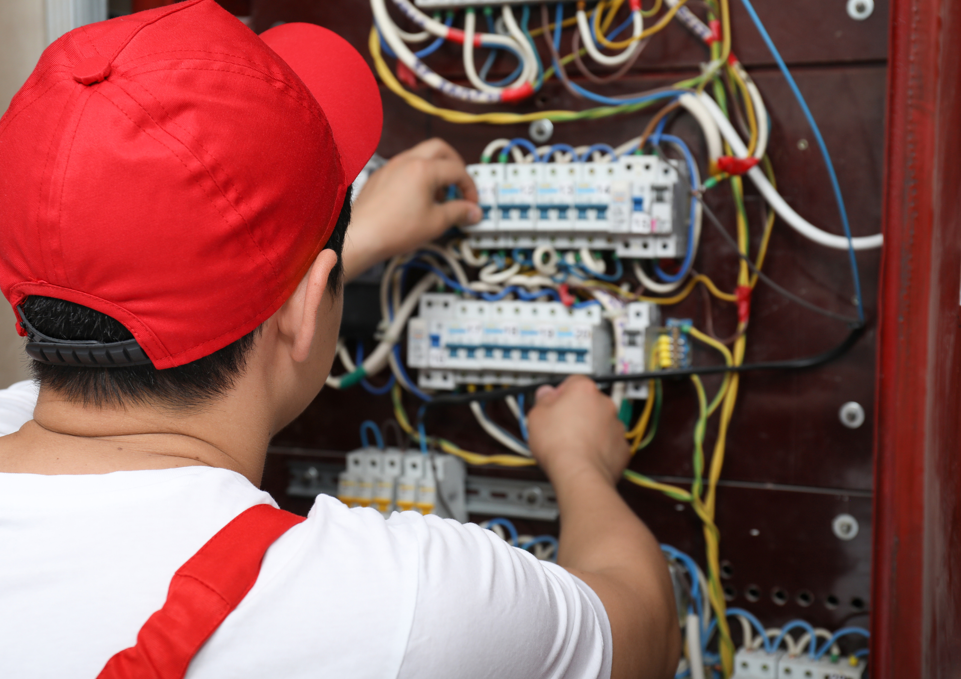 An electrician in a red cap and uniform works on electrical wiring inside a circuit breaker panel.