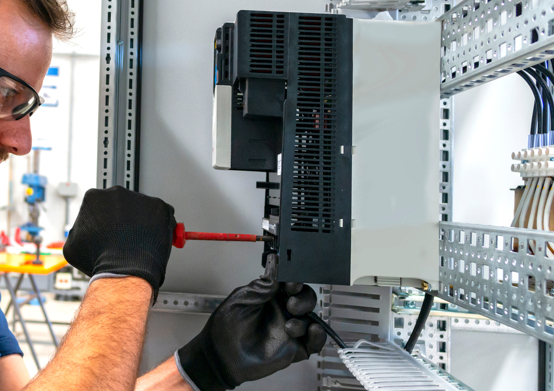 A technician wearing protective glasses and gloves uses a screwdriver on an industrial electronic device in a control panel.