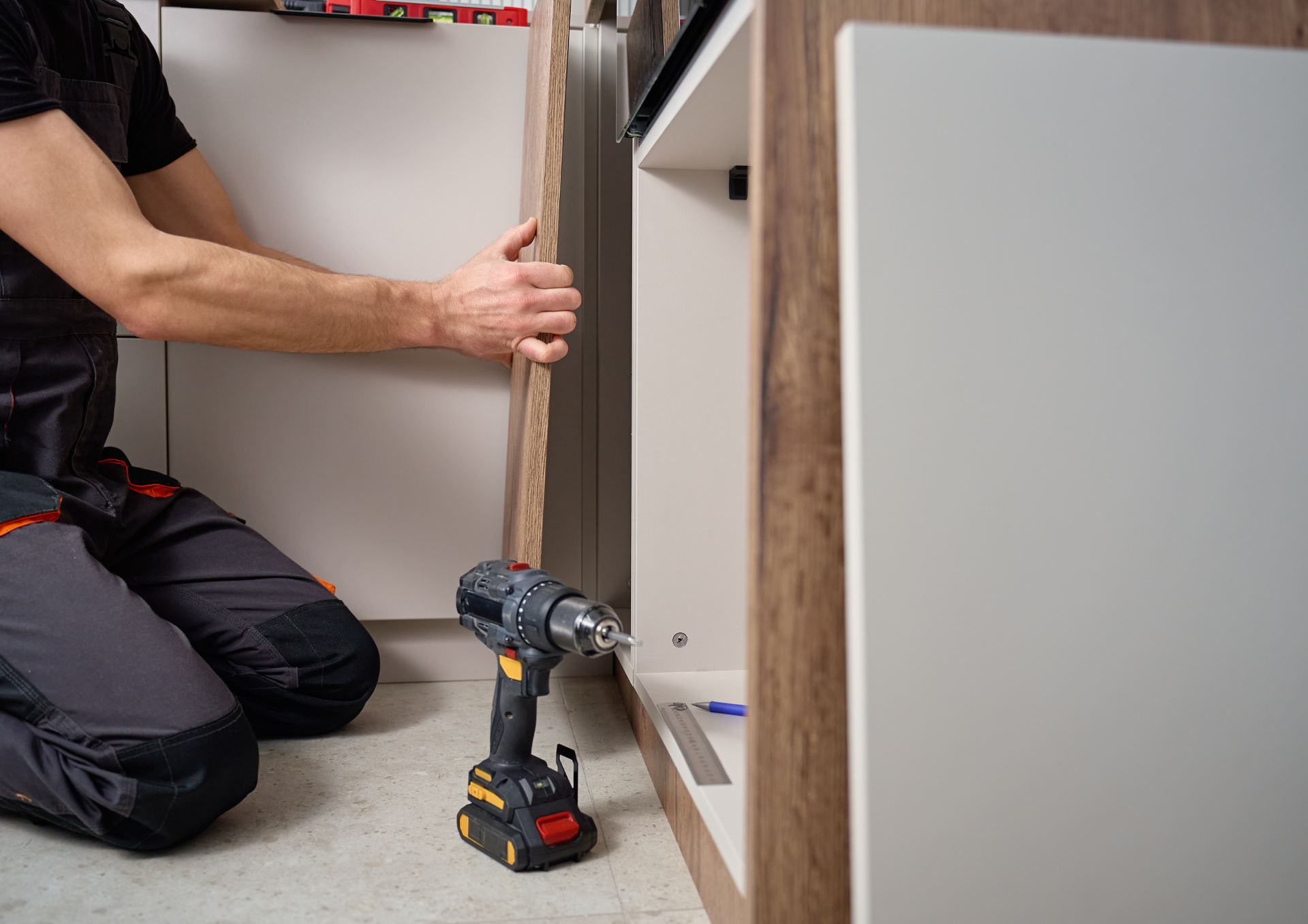 A person kneeling while installing a cabinet door with a cordless drill in a kitchen renovation setting.