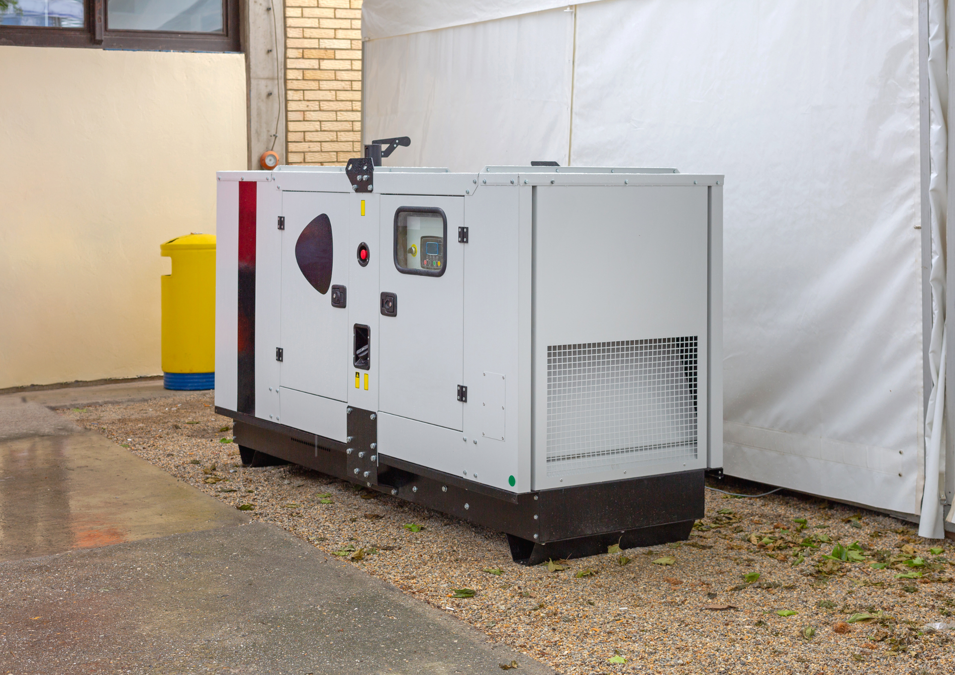A white industrial diesel generator sits on a gravel patch outside a building next to a white tent and yellow bollard.