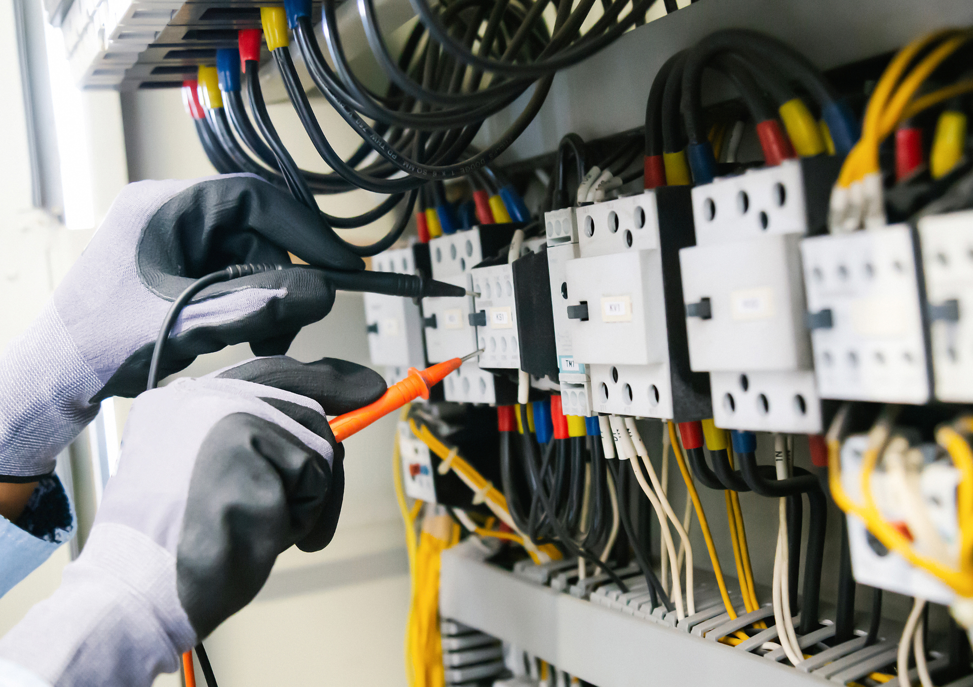 An electrician wearing protective gloves uses a multimeter to test electrical components inside a control panel.