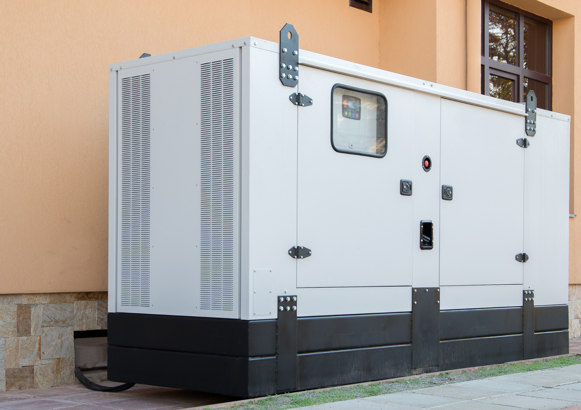 A large, white industrial generator sits outside a building with a tan wall and stone foundation.