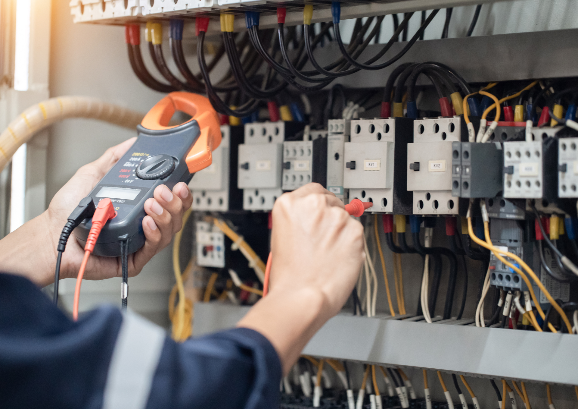 A technician uses a clamp meter and test lead to inspect electrical components inside a control panel.