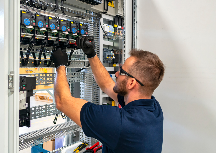 A technician wearing safety glasses and black gloves wires an electrical control cabinet.