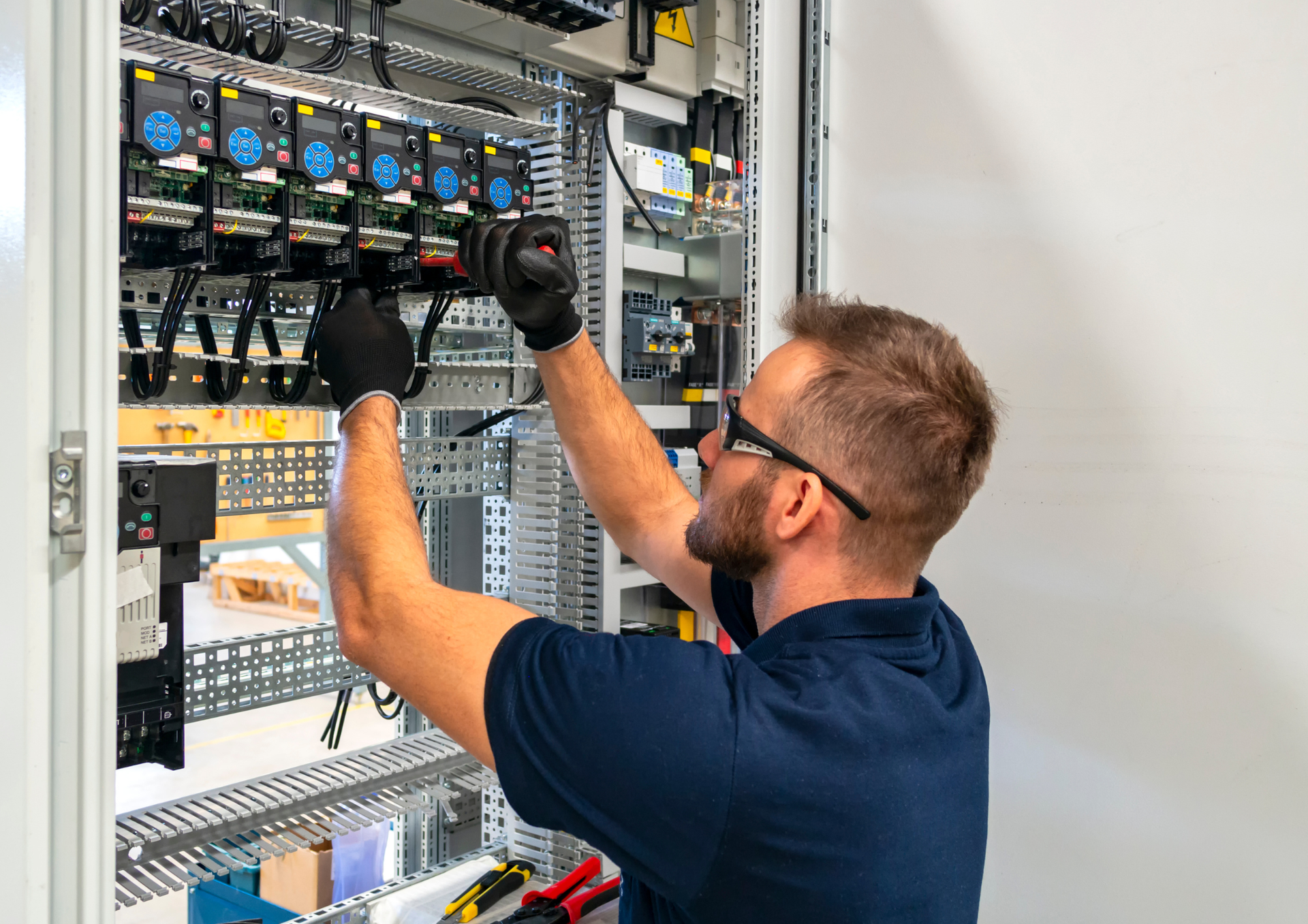 A person in a blue shirt and safety glasses works on wiring inside an electrical control panel.