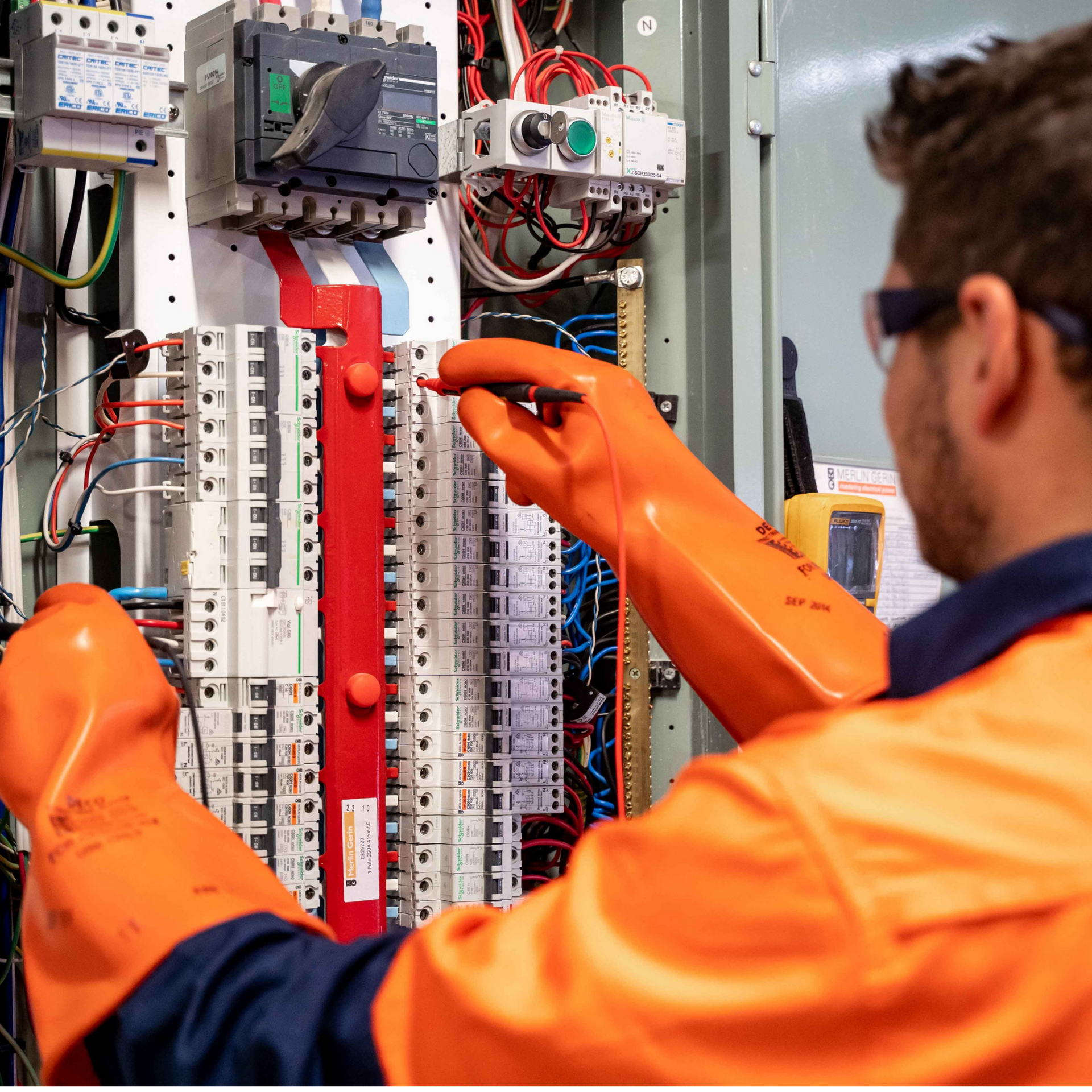 An electrician wearing orange safety gloves and high-visibility clothing tests the electrical components in a panel.