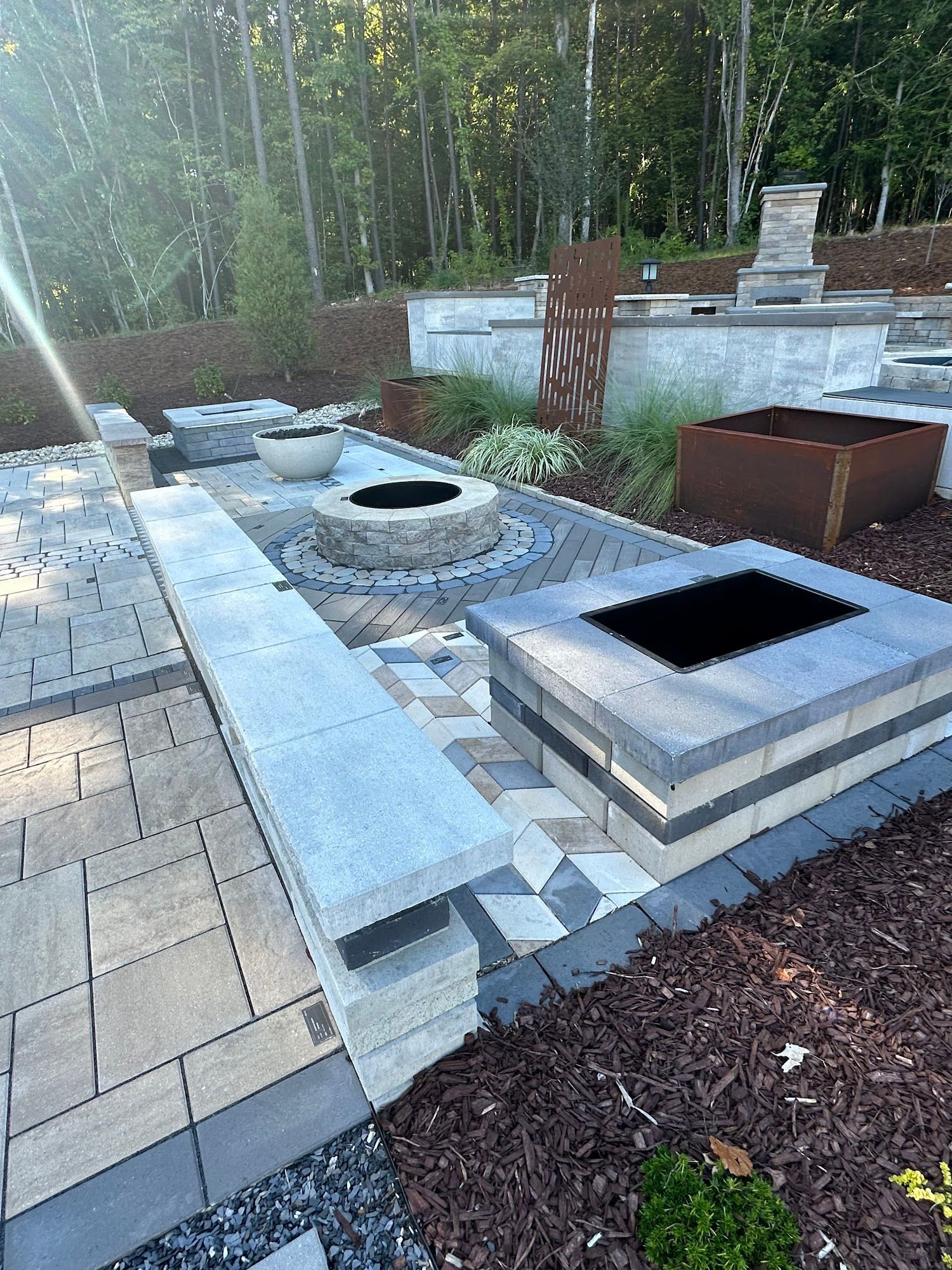 Stone patio with built-in fire pits, seating, and landscaping in a wooded setting.