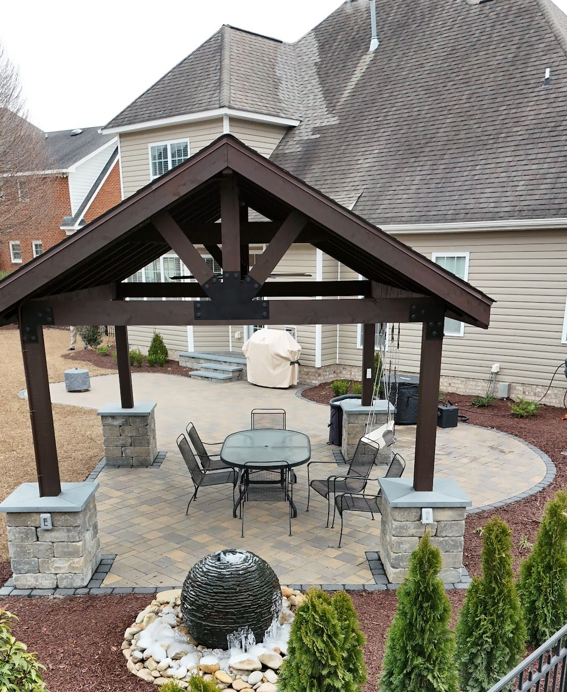 Patio with gazebo, stone pillars, furniture, and a water feature in a backyard.