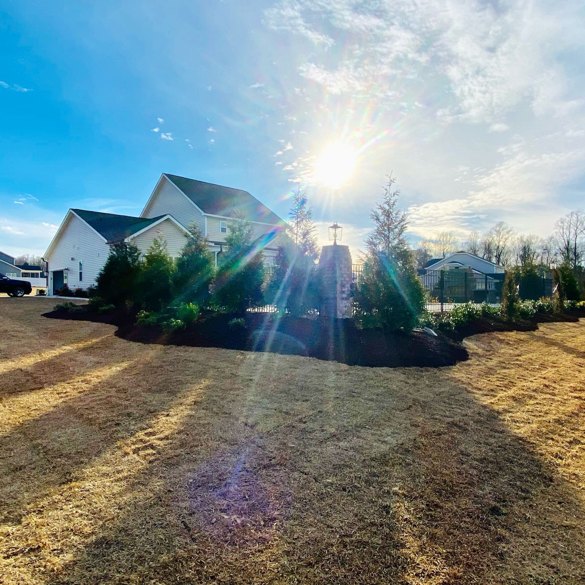 Sunlit house with landscaping: brown lawn, mulched beds, evergreen shrubs, and bright sky.
