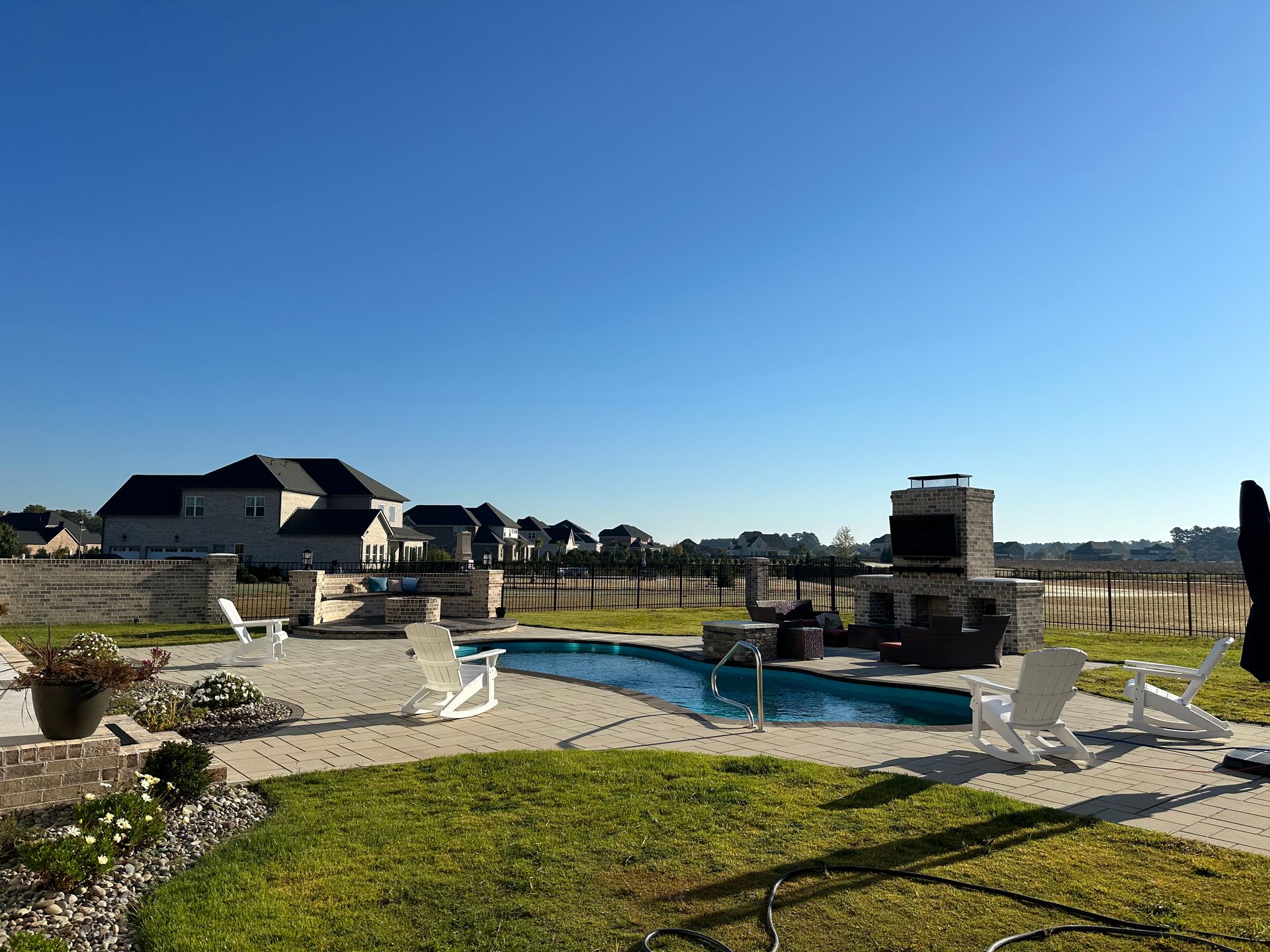 Backyard with pool, outdoor fireplace, white chairs, and houses under a clear blue sky.