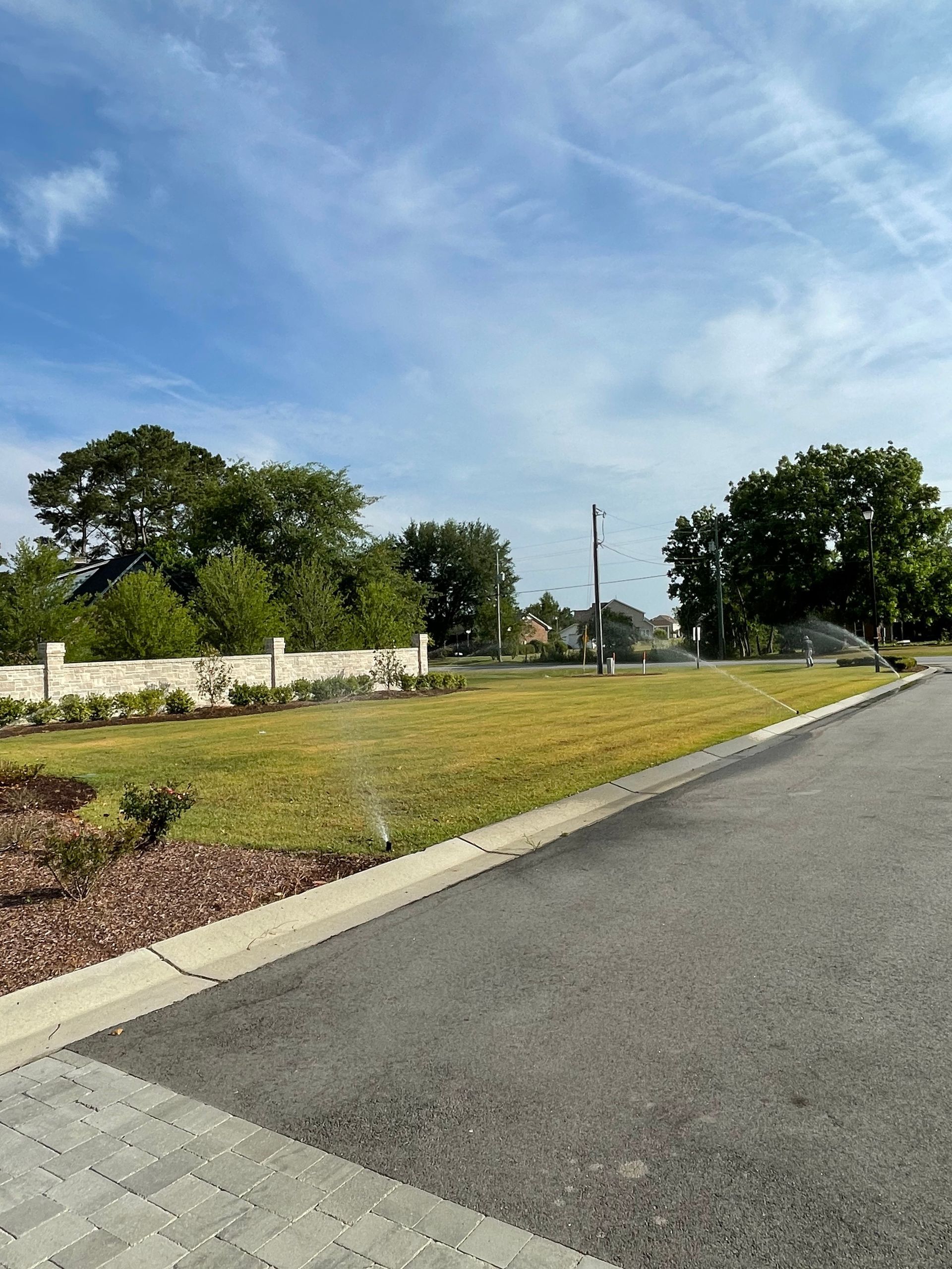 Lawn sprinkler watering grass next to a paved road and stone wall on a sunny day.