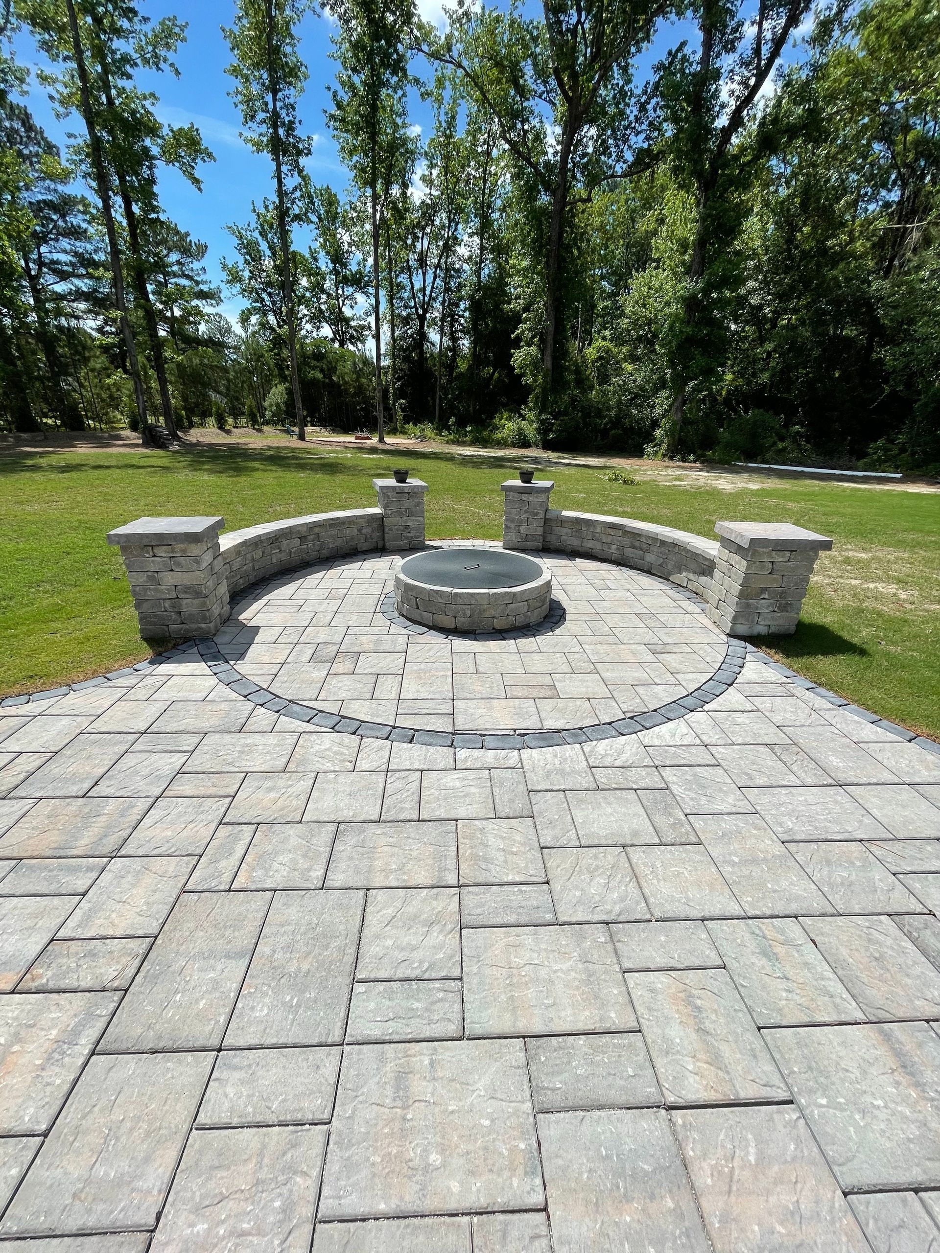 Stone patio with fire pit, seating, and pillars. Green grass and trees in the background under a blue sky.