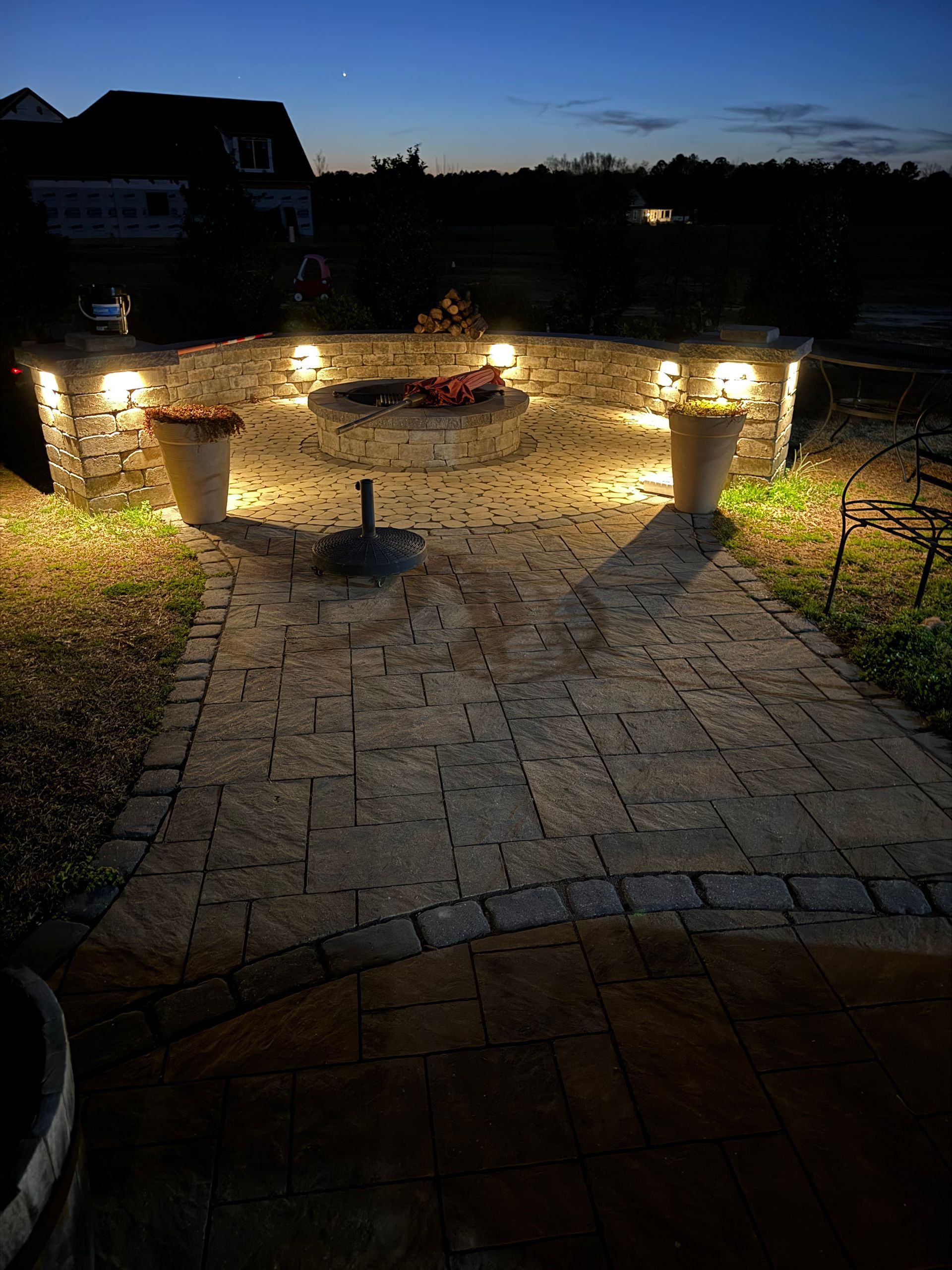 Lit stone patio with fire pit, planters, and wall lights at dusk.