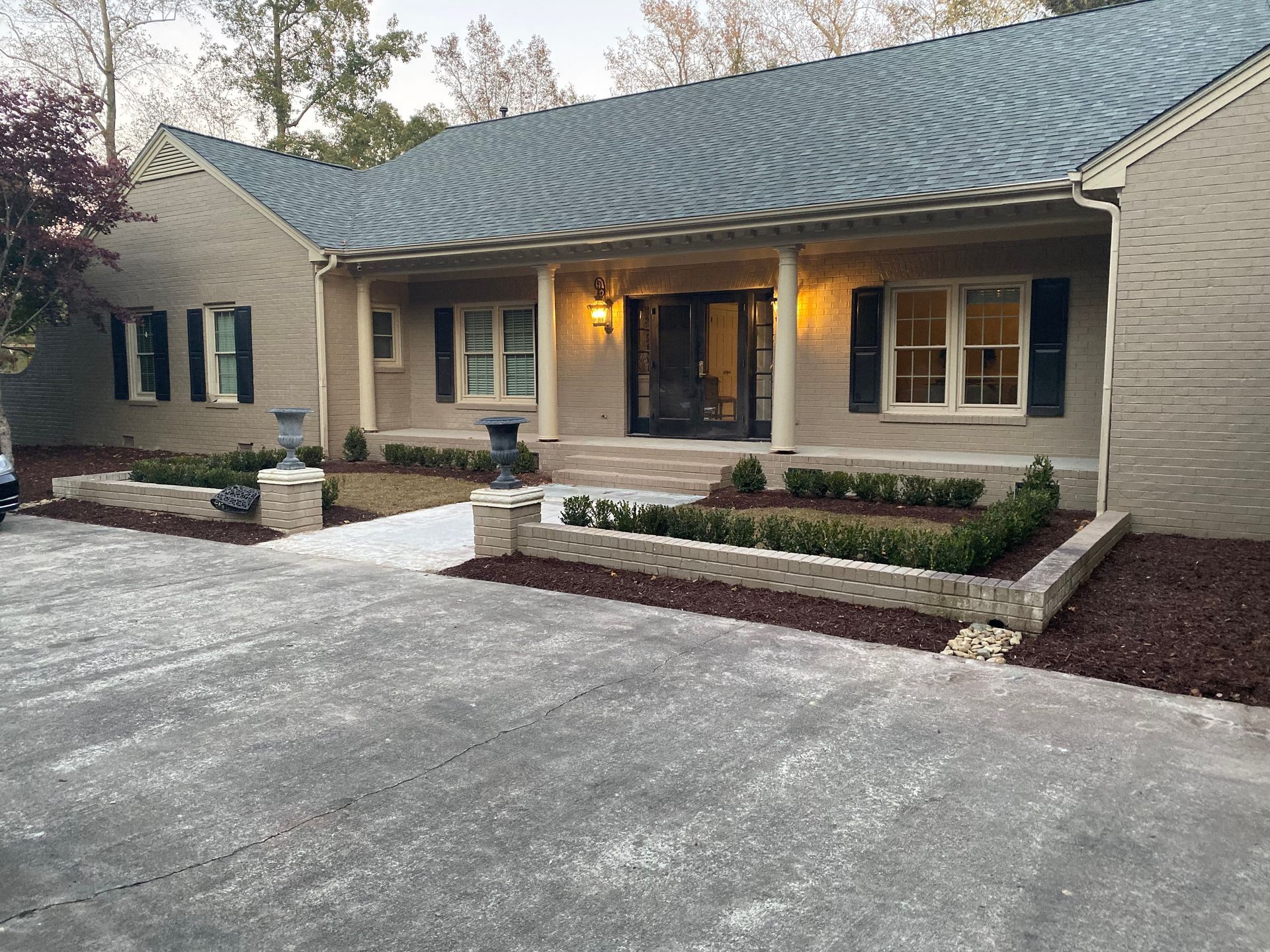 Grey brick home with black shutters, landscaping with mulch beds, and a concrete driveway.