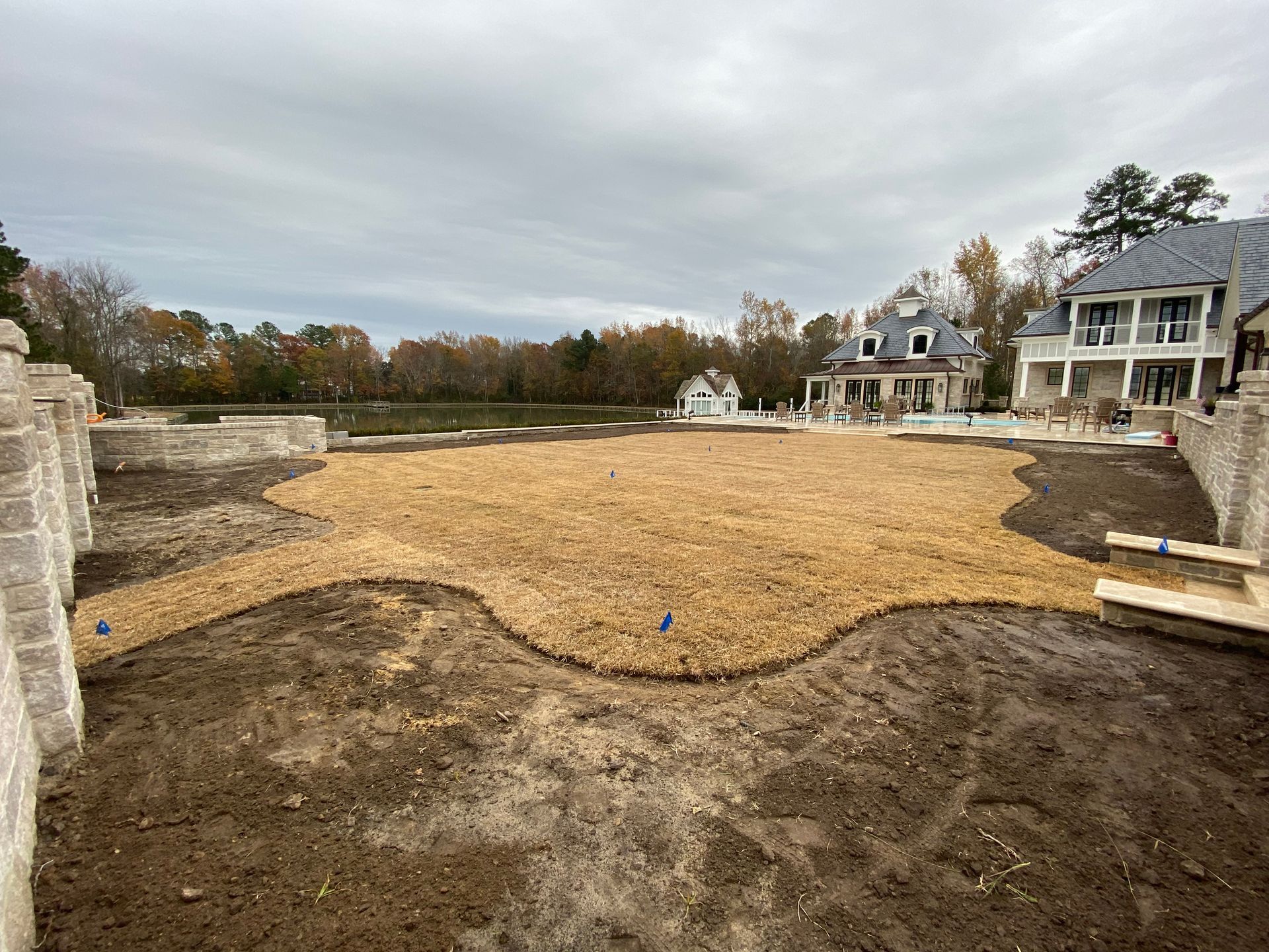 A large backyard with newly seeded grass, partially framed by stone walls, a house in the background, and cloudy sky.