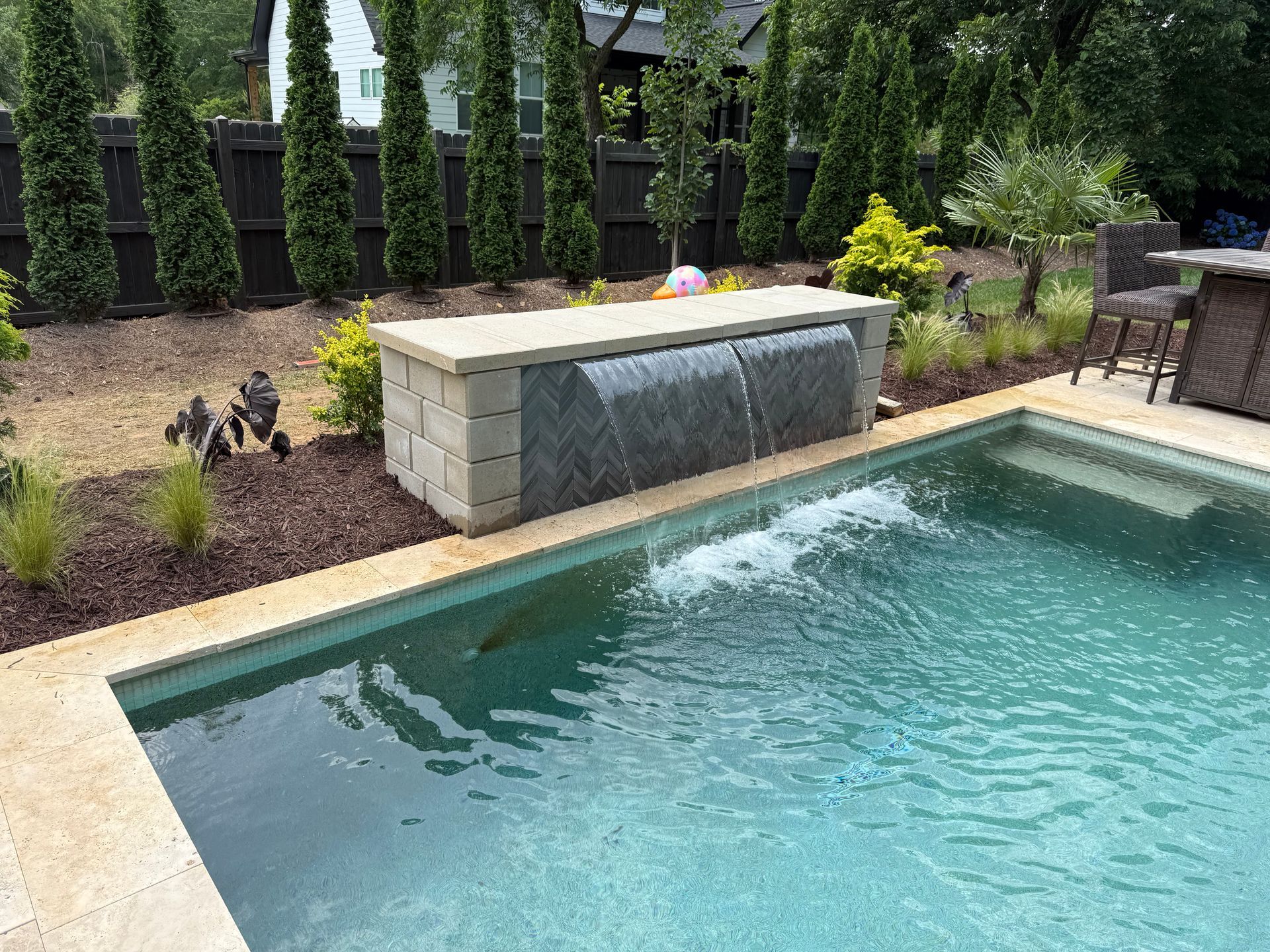 Pool with a waterfall feature; stone wall, turquoise water, landscaping and a brown fence.