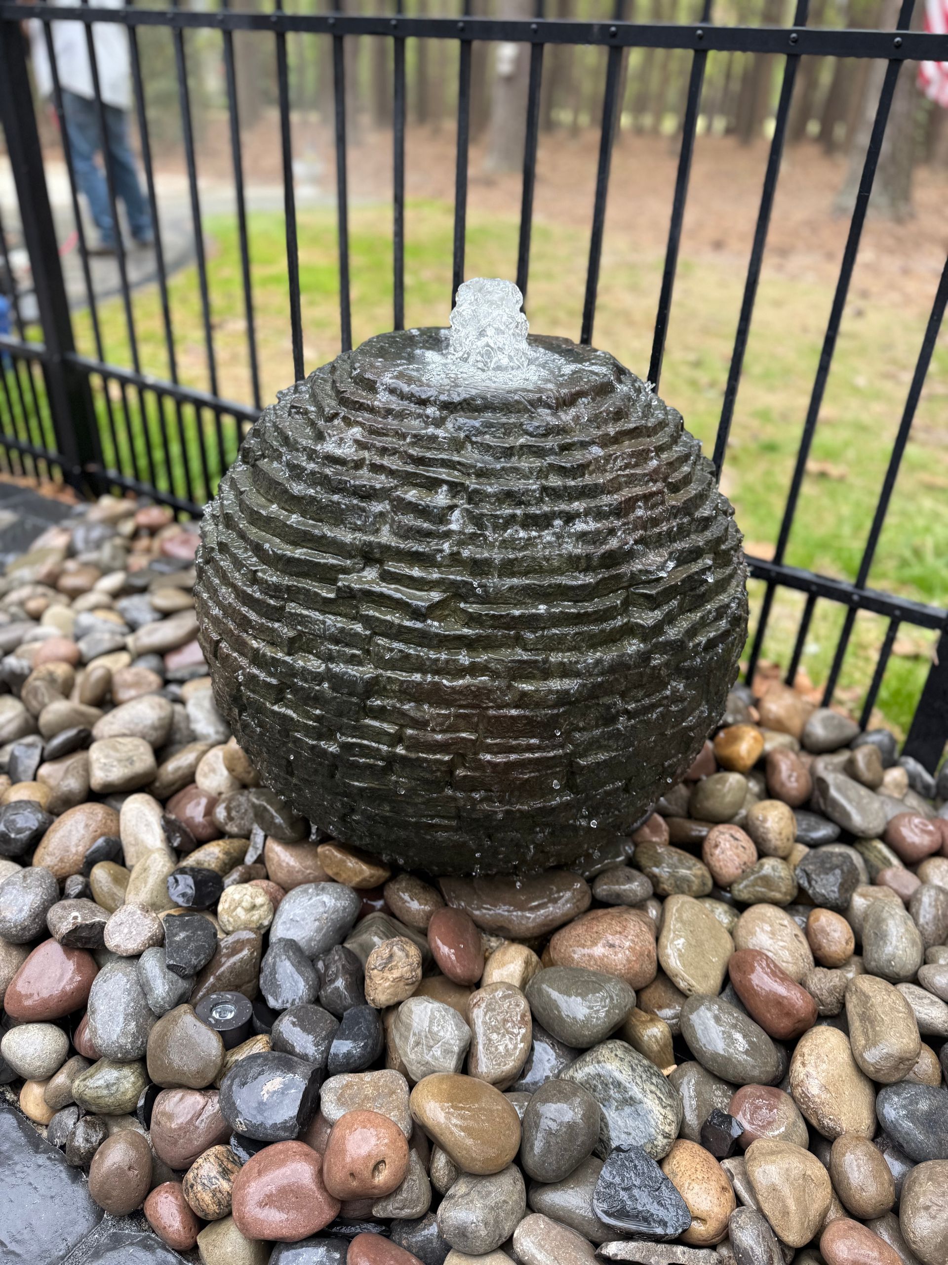 Spherical water fountain with water stream, surrounded by pebbles. Fence and trees in background.