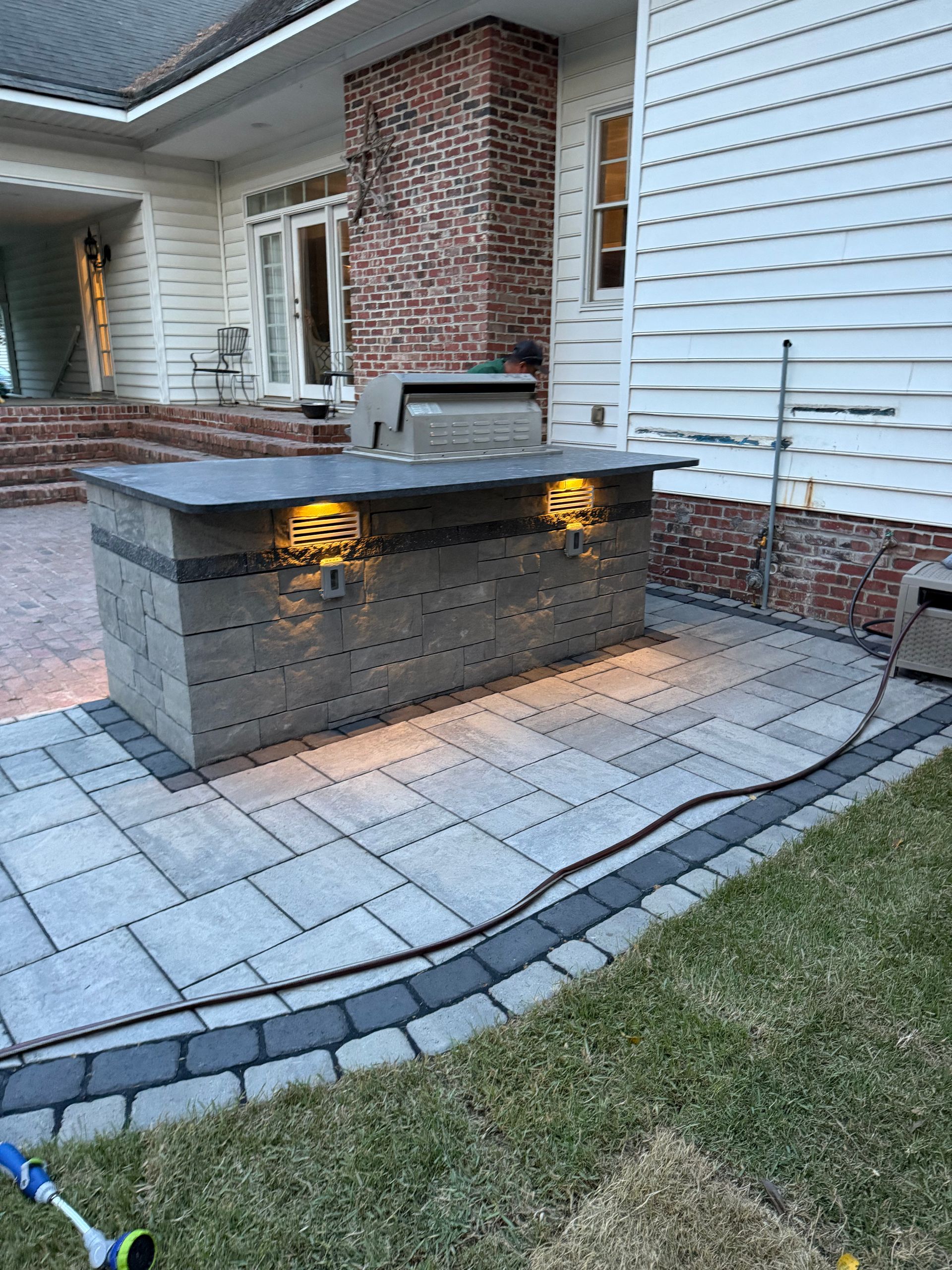 Outdoor stone patio with grill island, lighted, next to house.