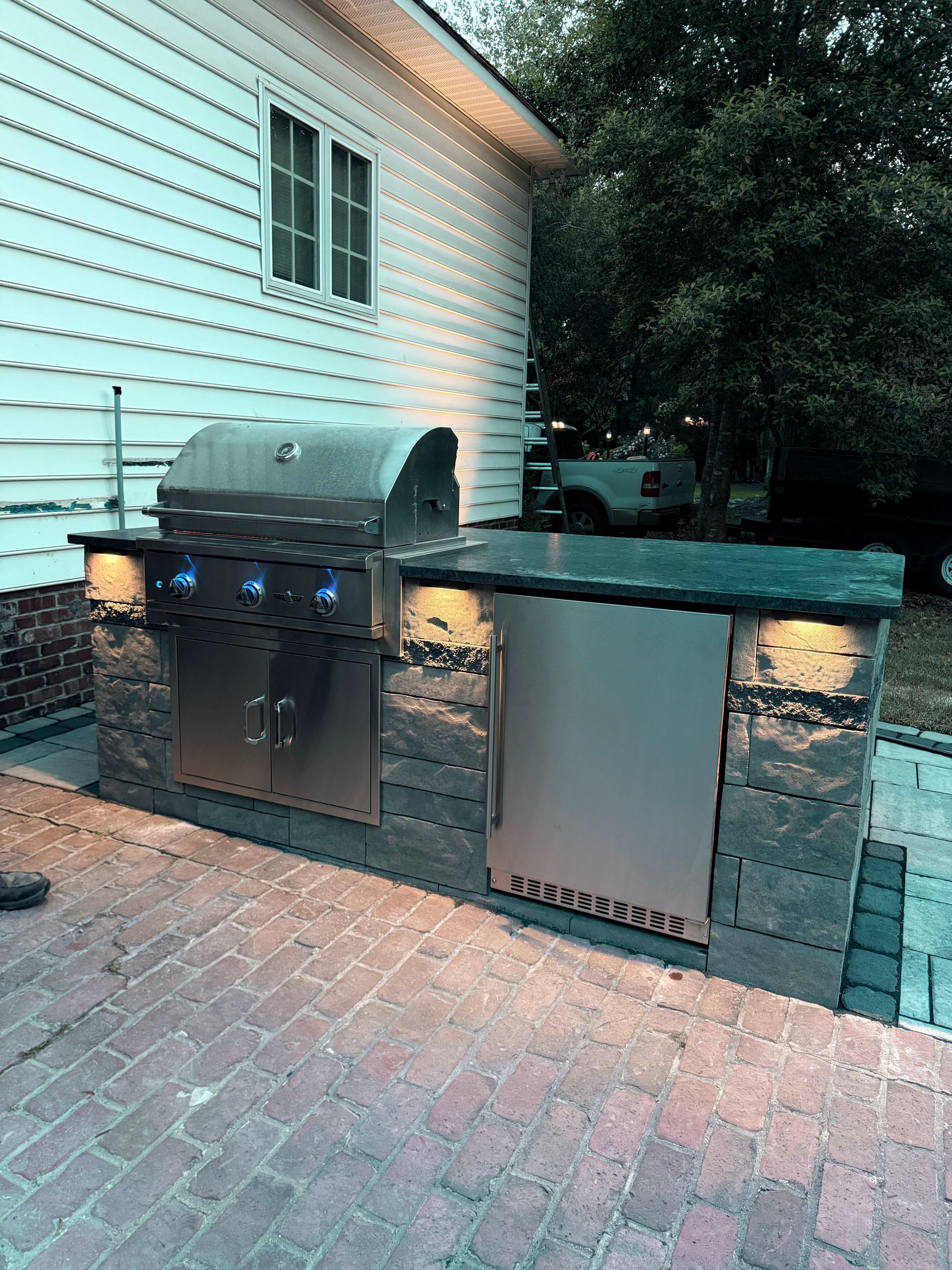 Outdoor kitchen with stainless steel grill, fridge, and stone surround under a house.