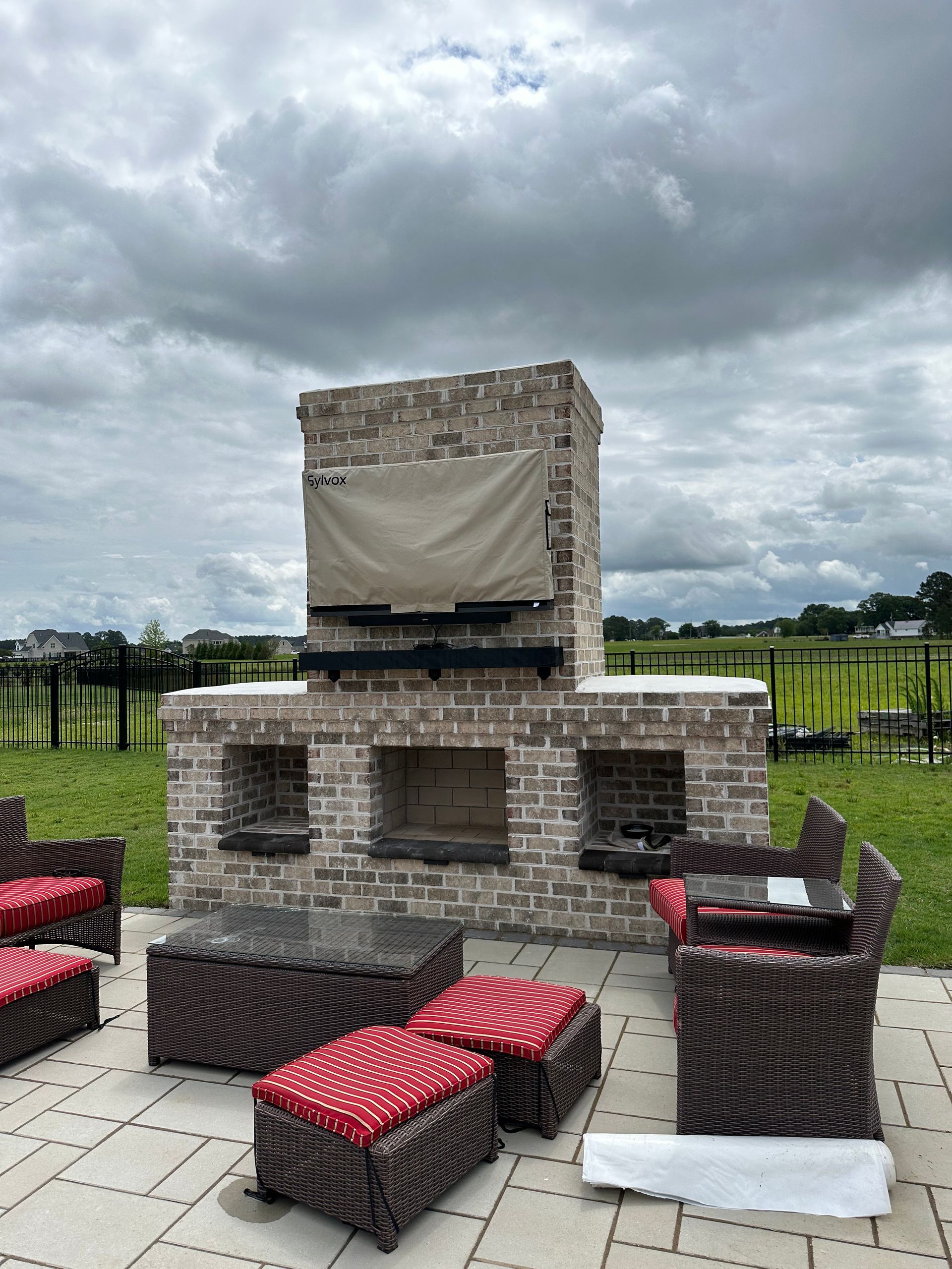 Outdoor brick fireplace with a covered TV, seating area on a patio, cloudy sky.