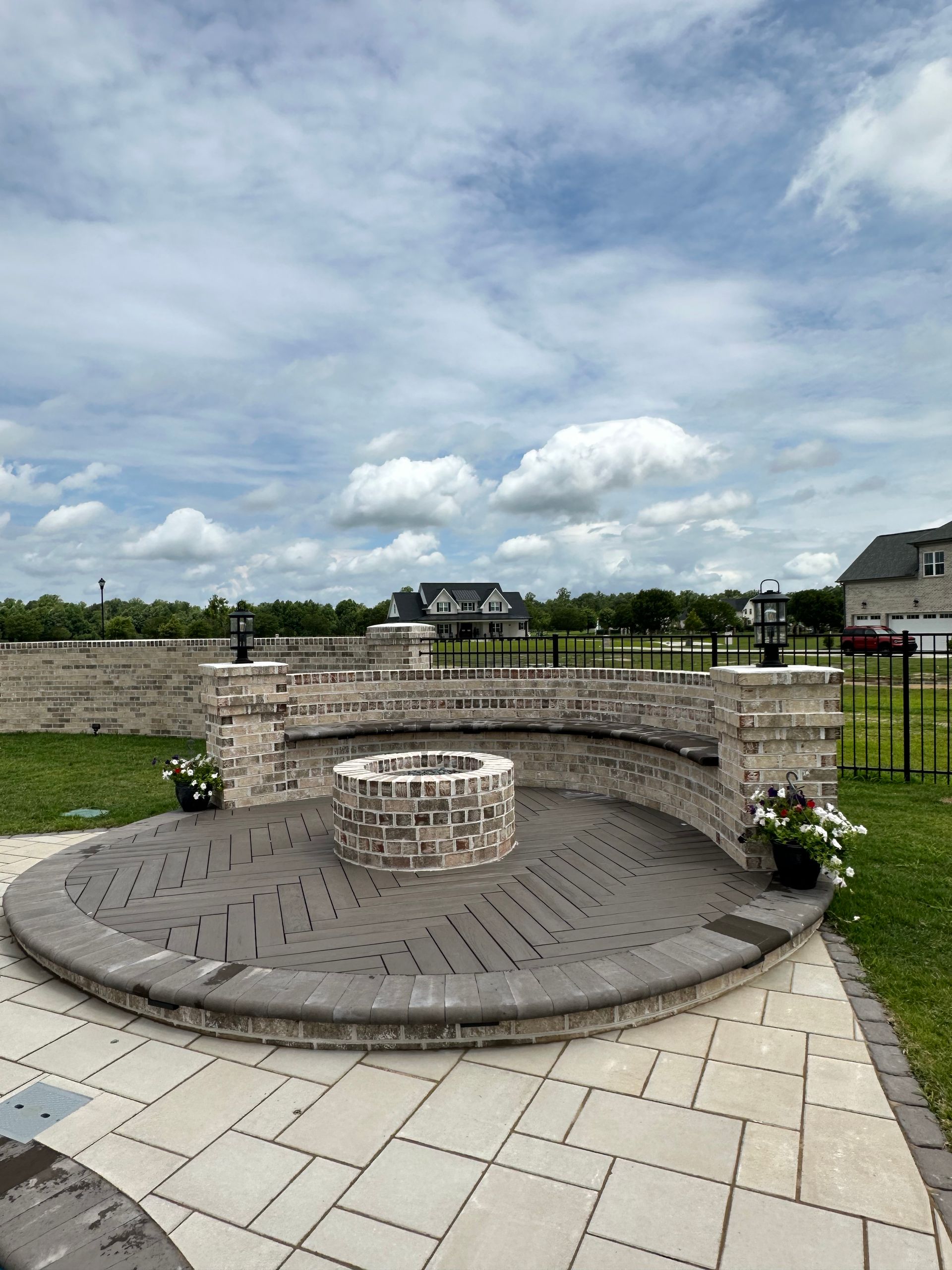 Brick fire pit area with seating, light brick wall, dark patterned ground, under cloudy sky.