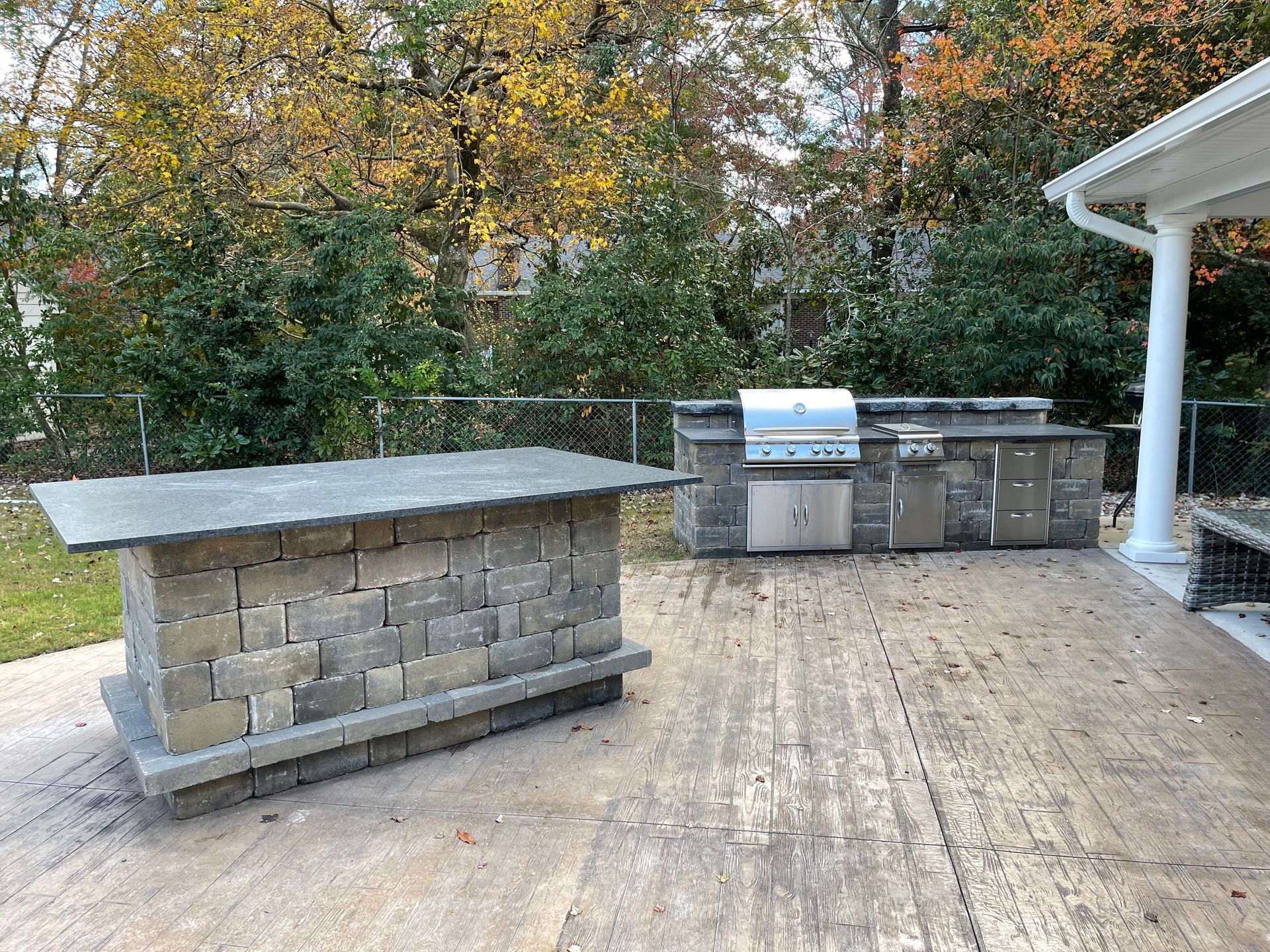 Outdoor kitchen with stone grill and bar on a wooden patio; trees in the background.