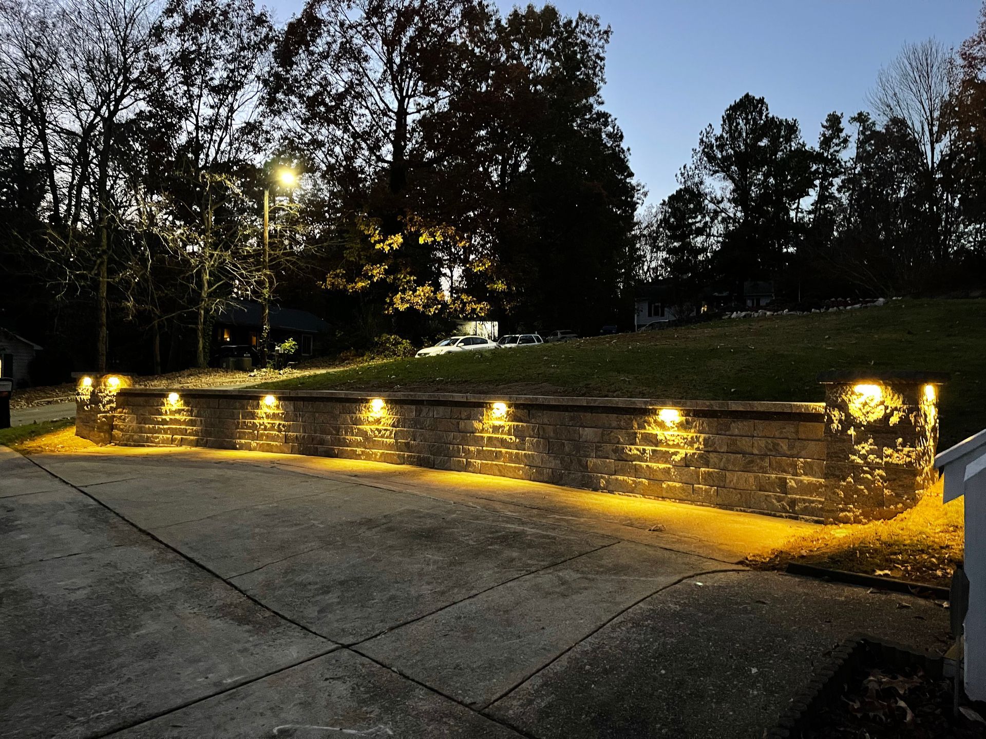 A retaining wall with built-in lights illuminates a dark driveway and lawn at dusk.