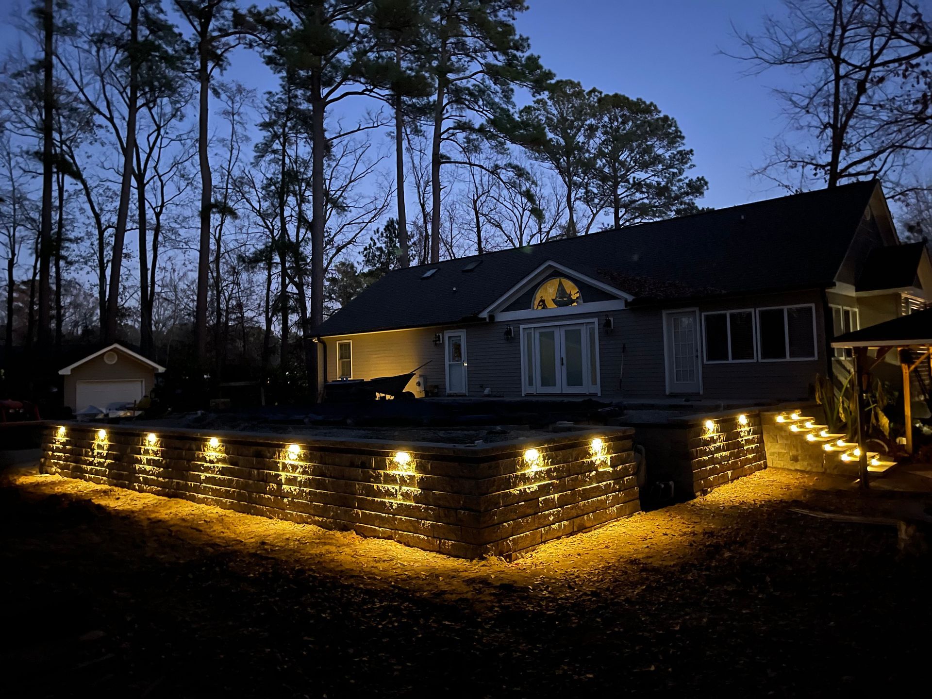 House exterior at dusk, illuminated by warm lights on retaining walls and steps. Trees in background.