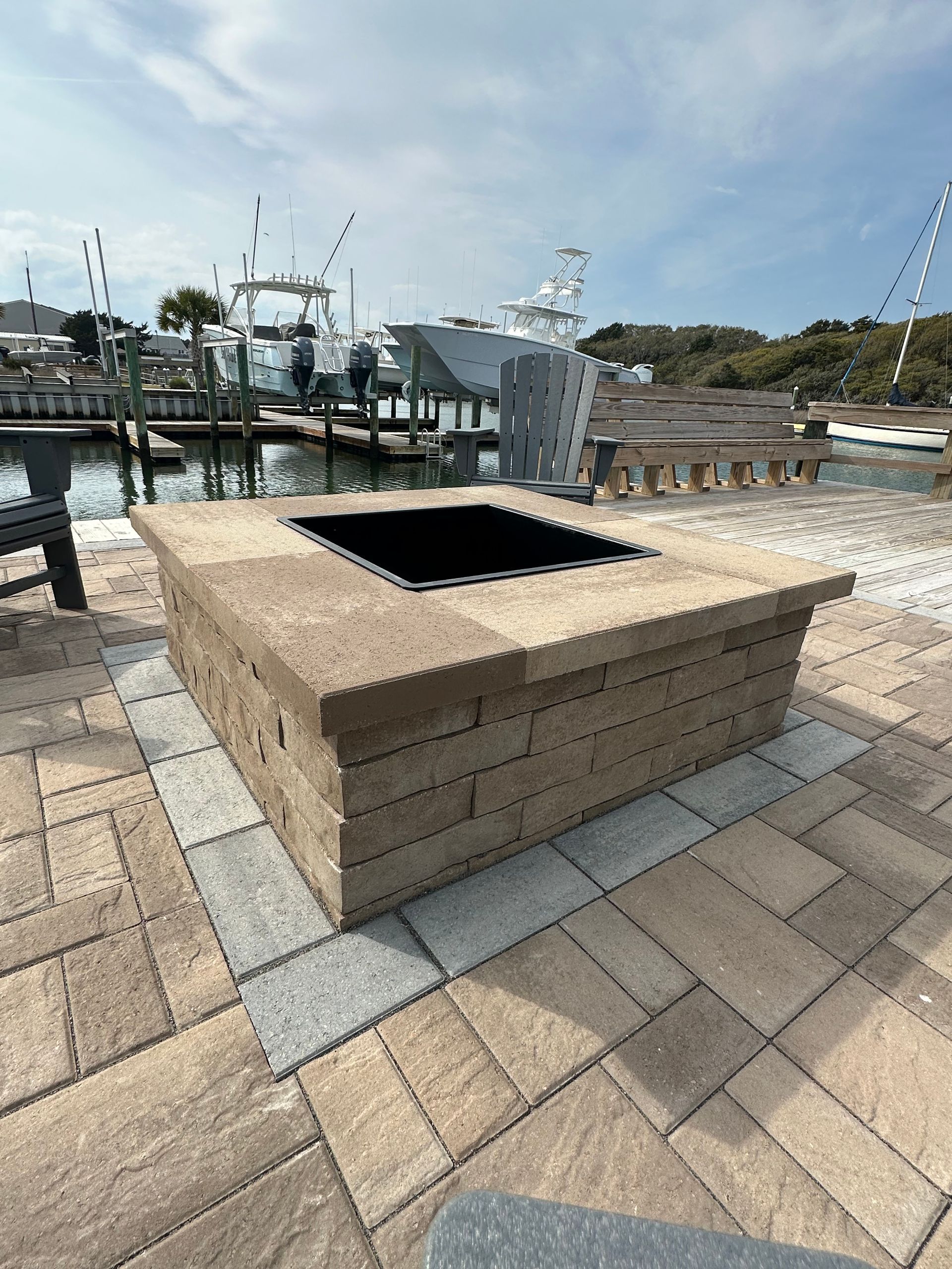 Stone fire pit on a brick patio by a marina with boats and benches.